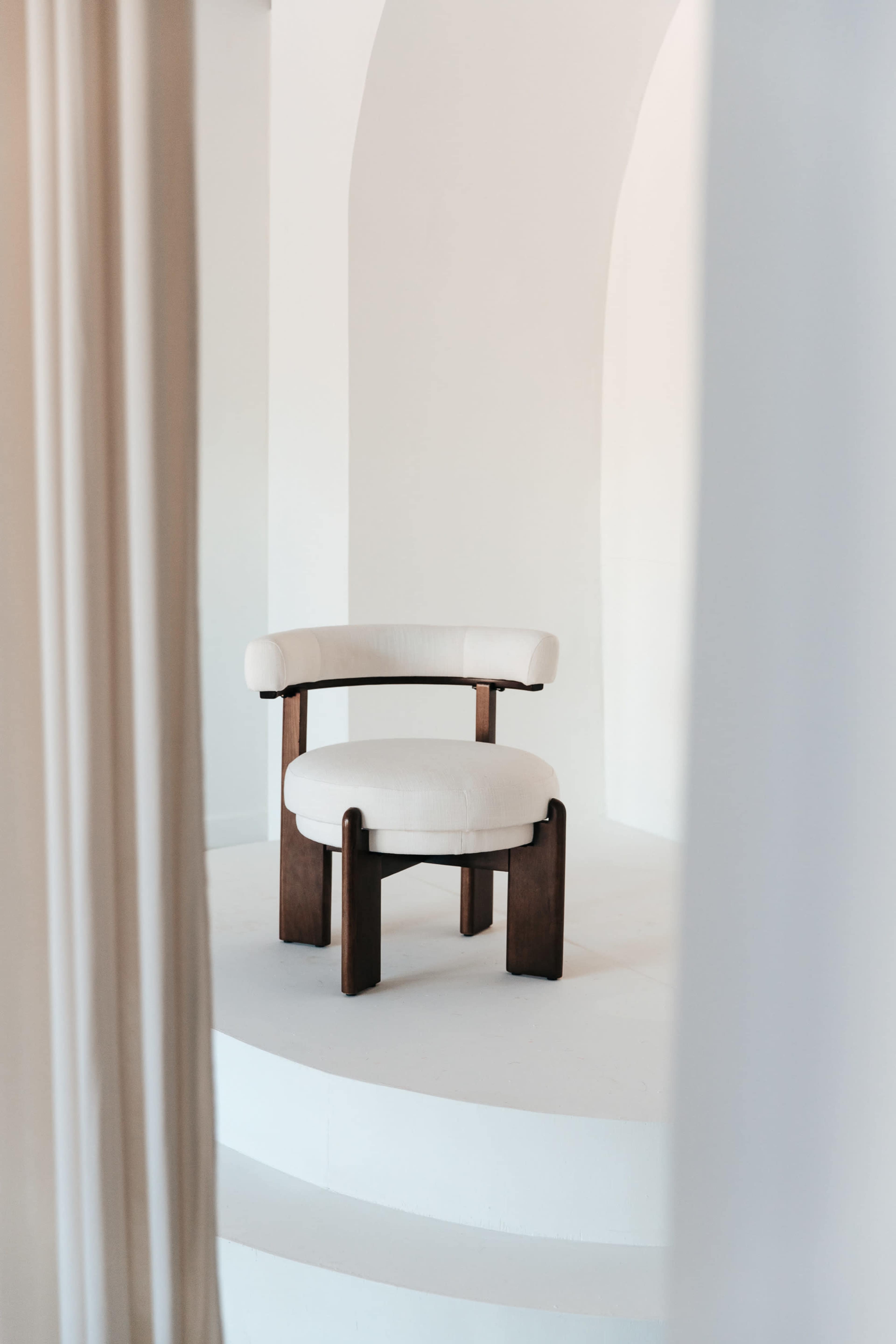 A modern, off-white upholstered chair is placed on a circular white platform against a minimalist backdrop.