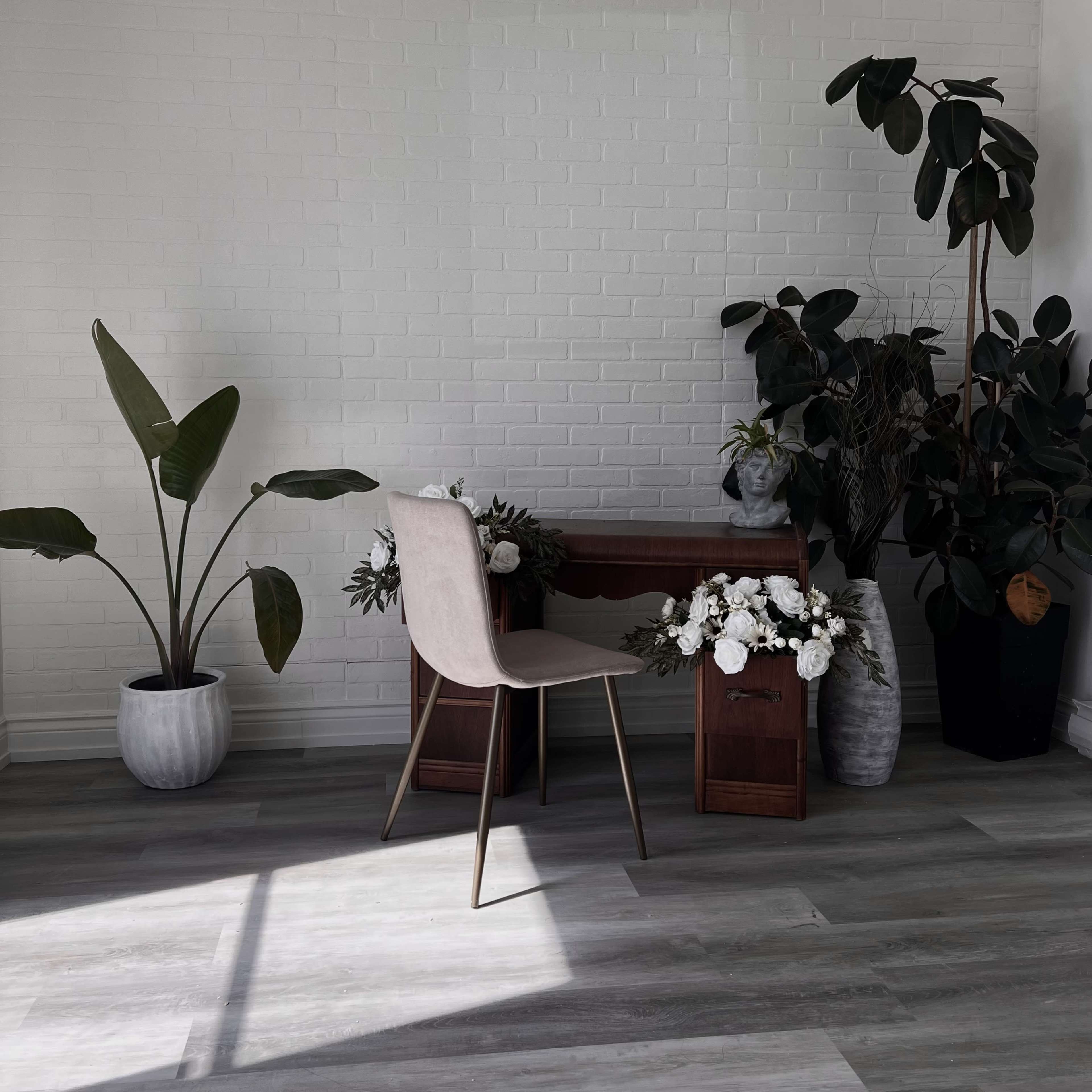 A minimalist workspace features a chair and desk surrounded by plants and floral arrangements against a white brick wall.