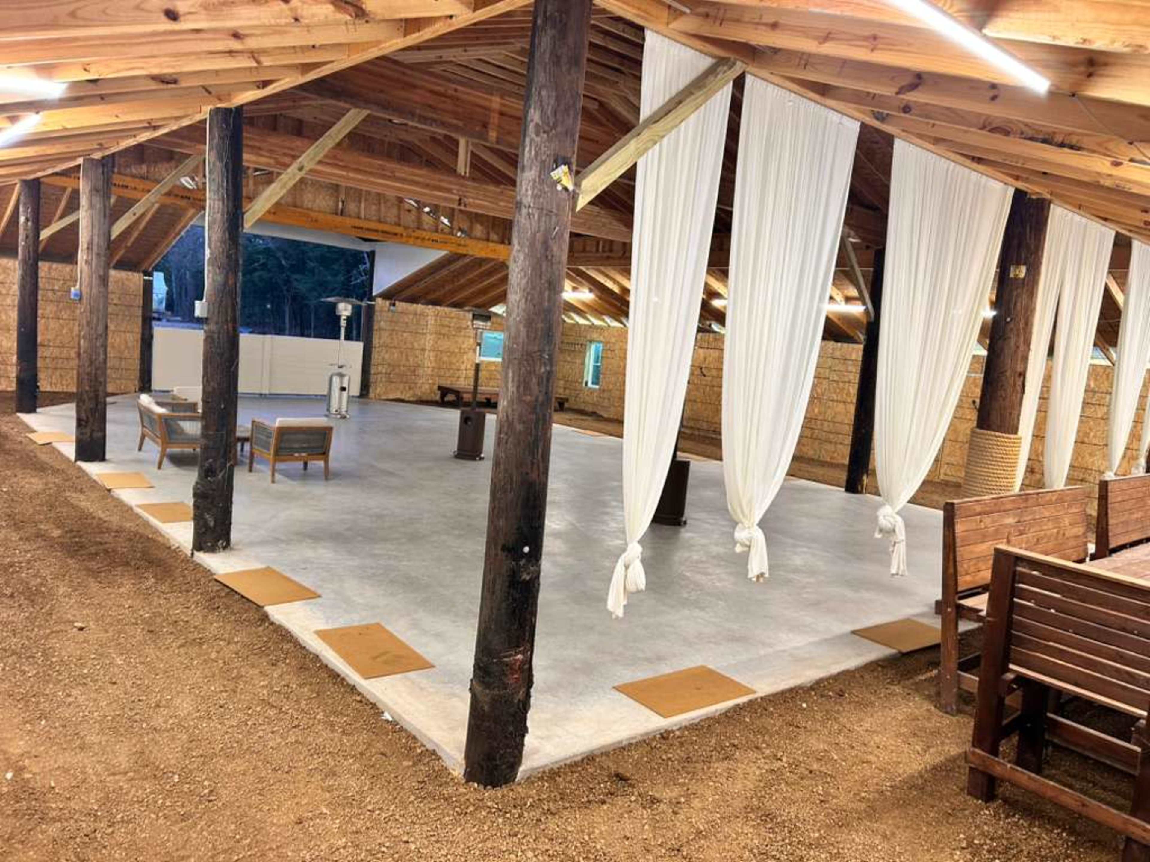 The image shows a spacious indoor area with a polished concrete floor, wooden beams, and white drapes hanging from the ceiling, surrounded by natural light.