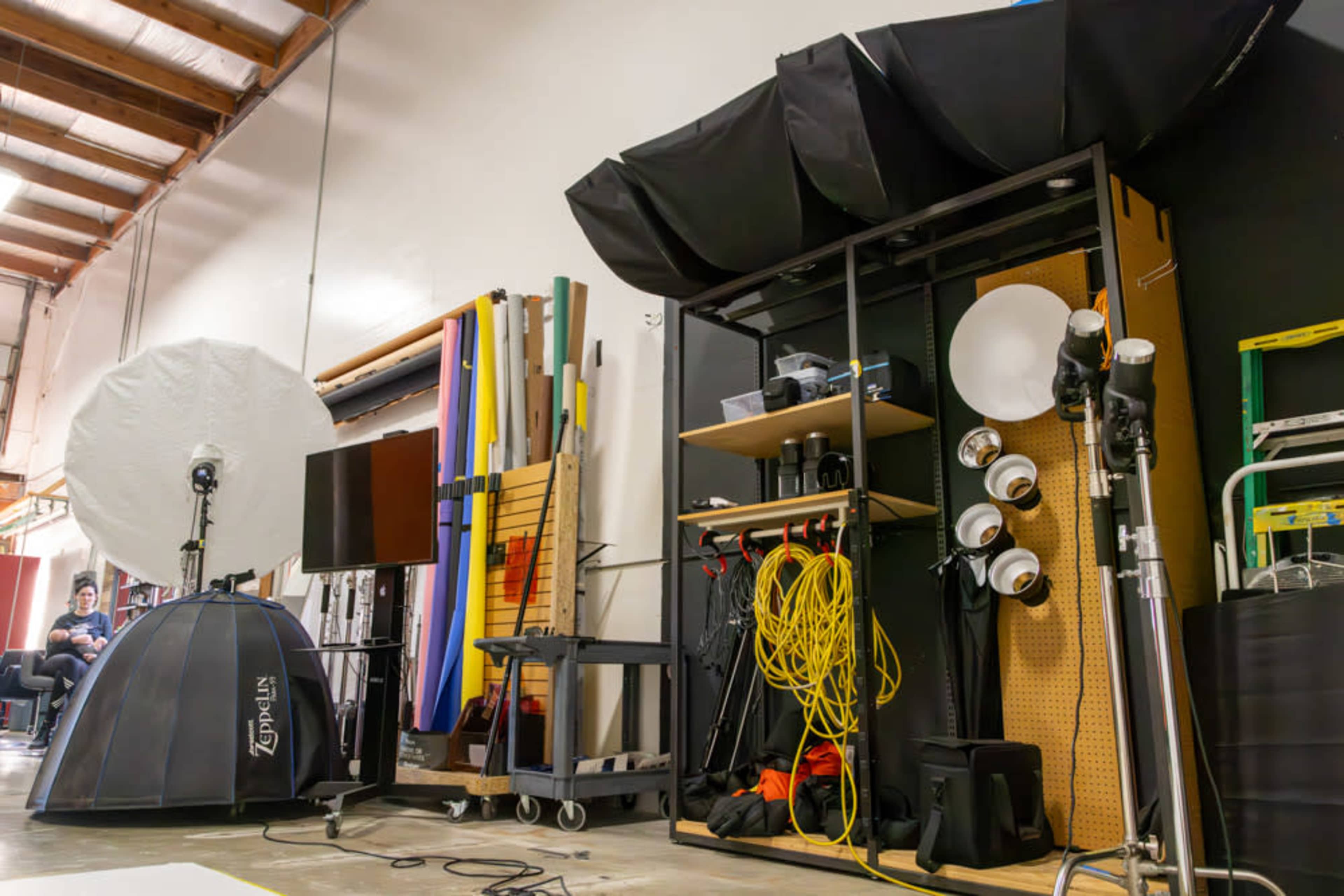 The image shows a photography studio setup with various lighting equipment, backdrops, and storage carts along a wall.