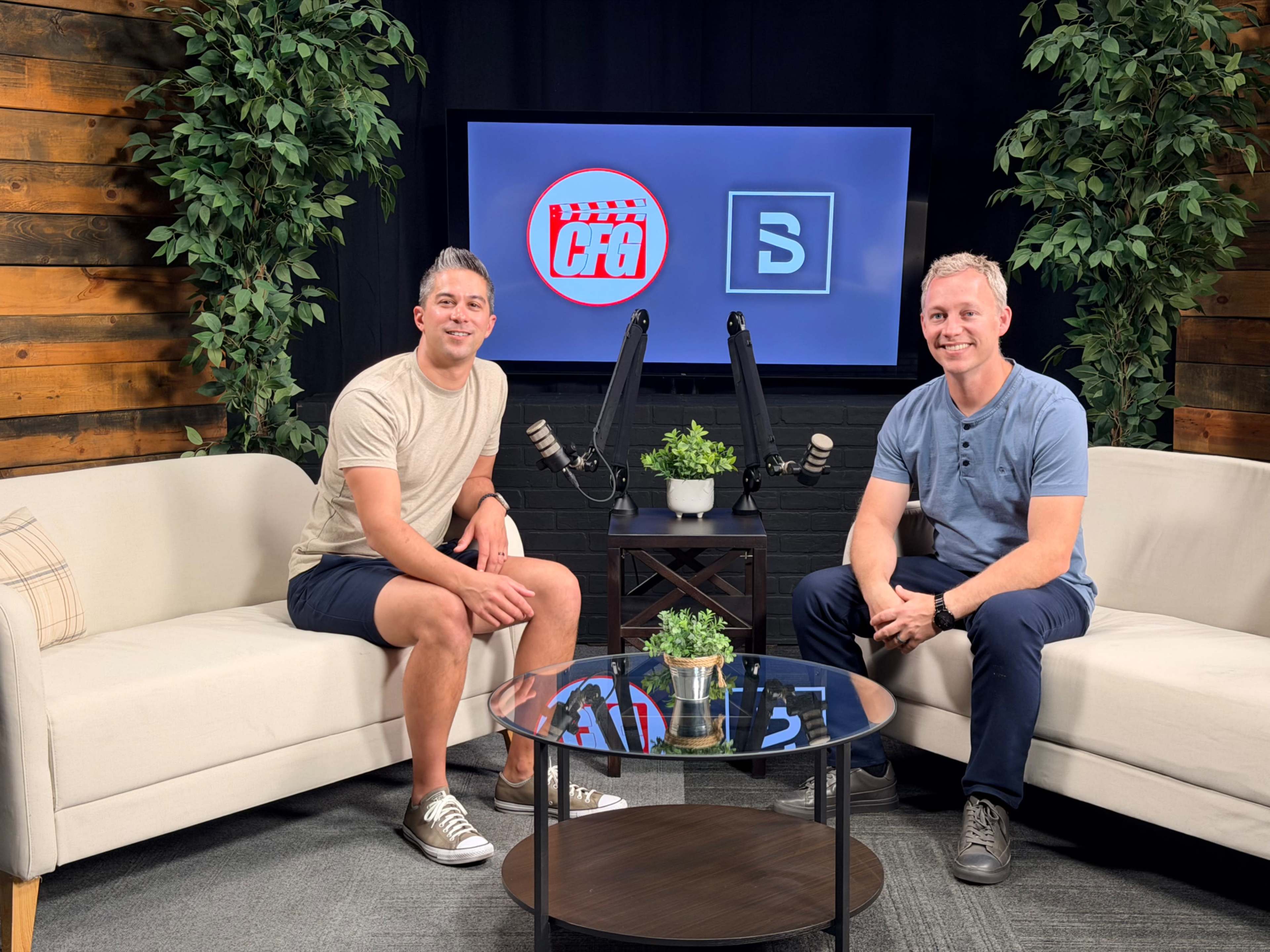 Two men are seated on a couch in a studio set, facing the camera with a table and plants in front of them, and screens displaying logos in the background.