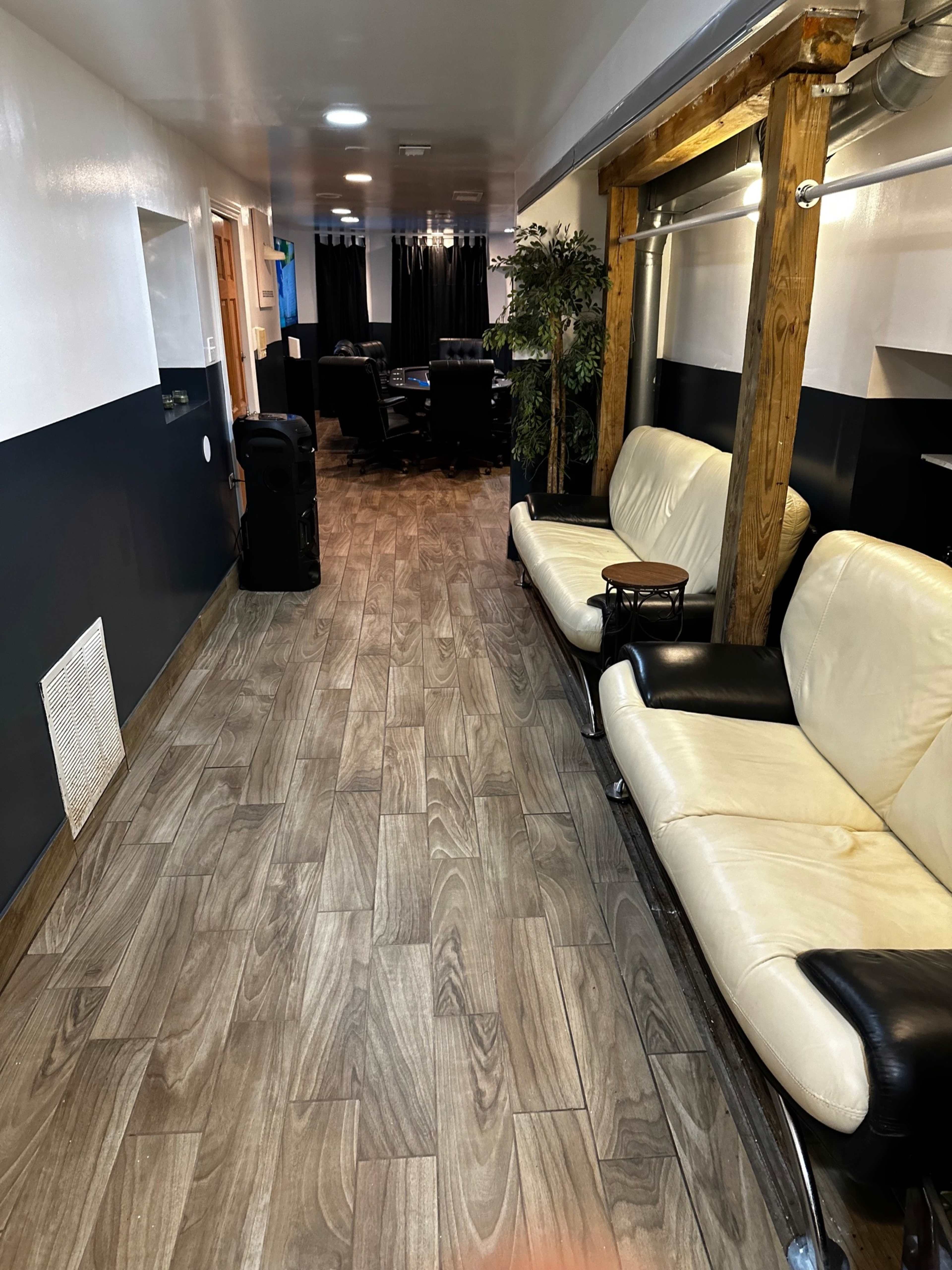 The image depicts a long hallway featuring wooden flooring, two couches on one side, and a view of a meeting room at the end.