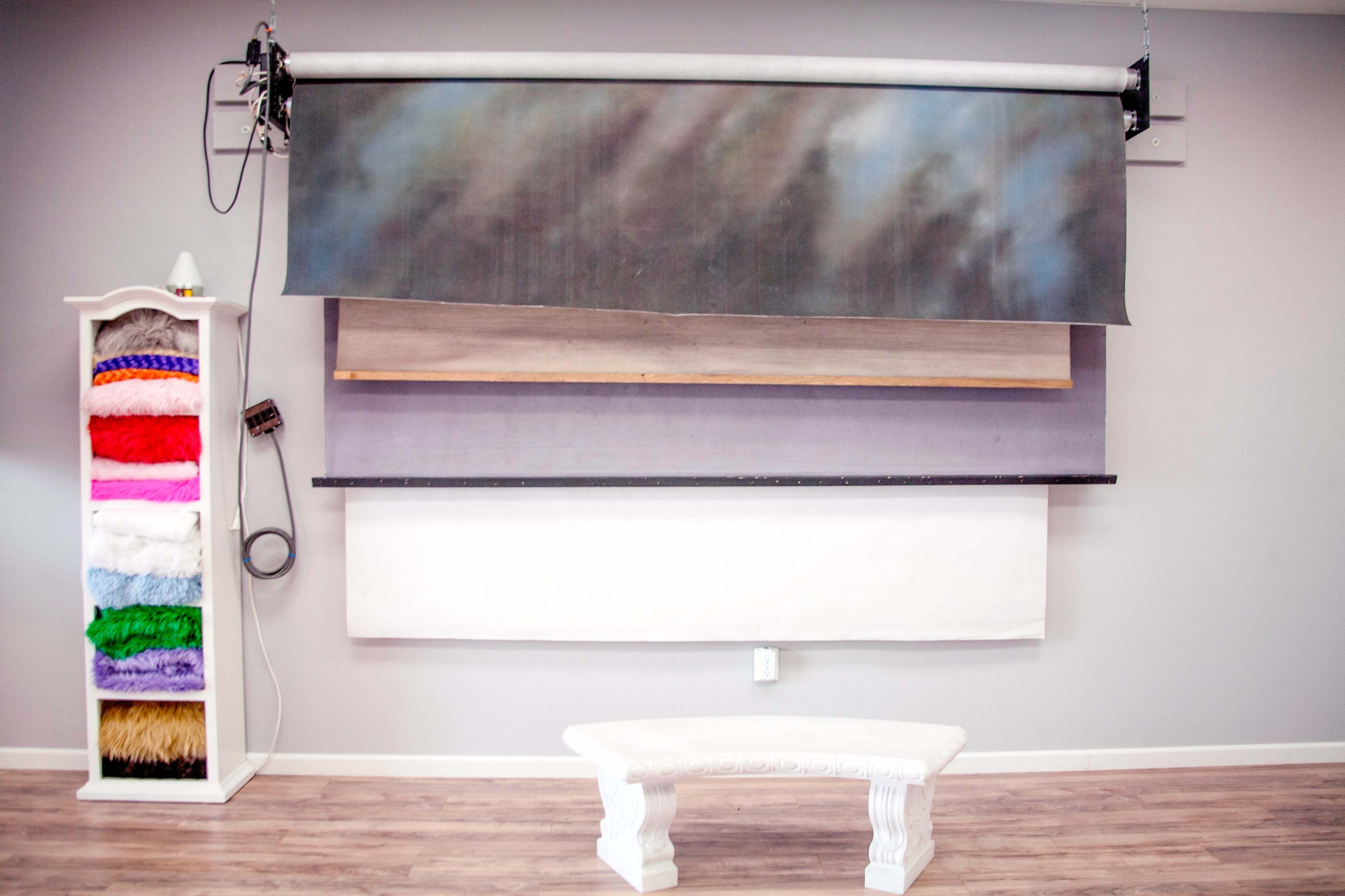 A photography studio setup with a backdrop system and a white bench in front of a wall displaying various colored fabric rolls.
