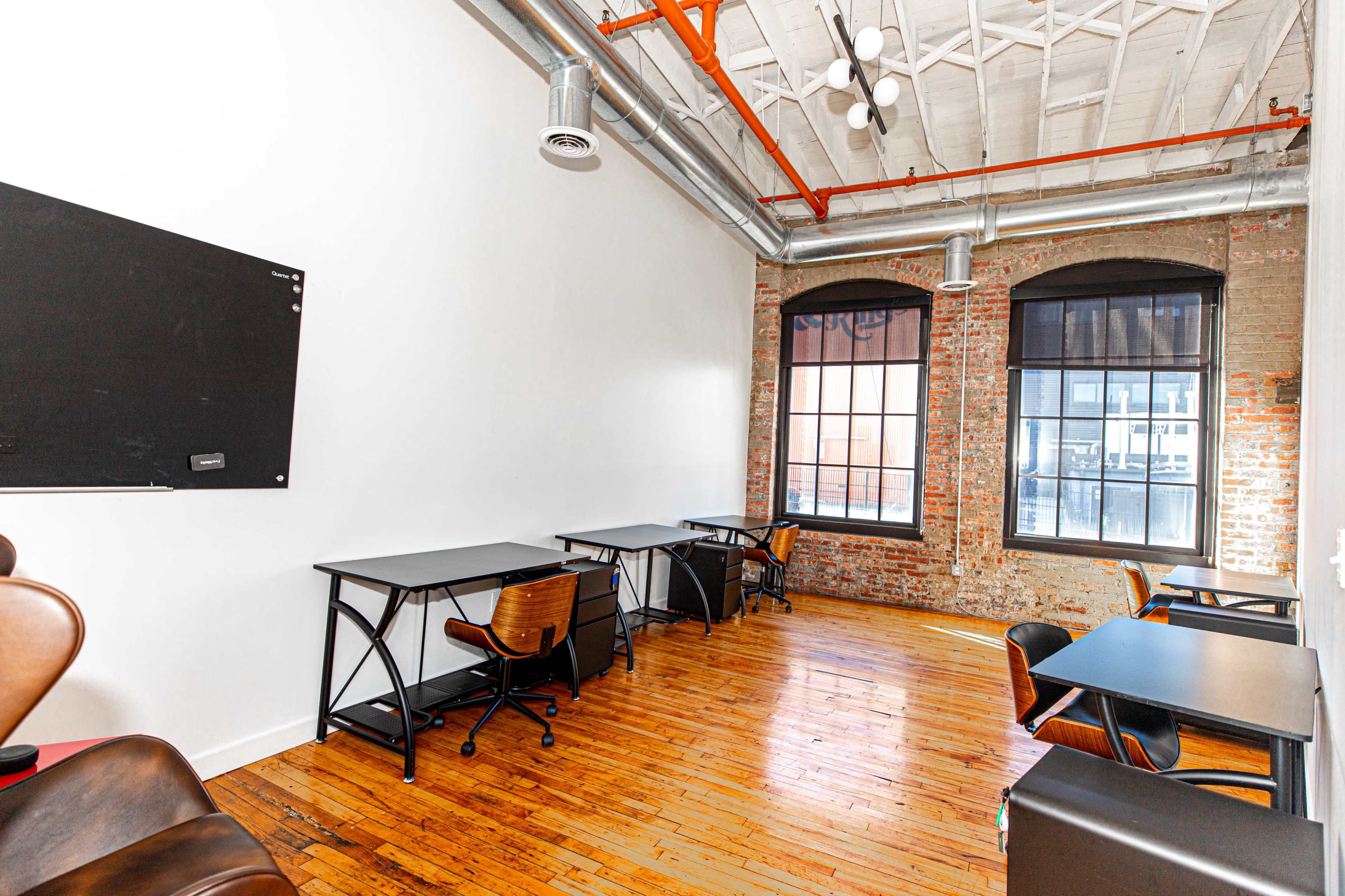 A spacious office with wooden floors, exposed brick walls, and several desks arranged near large windows.