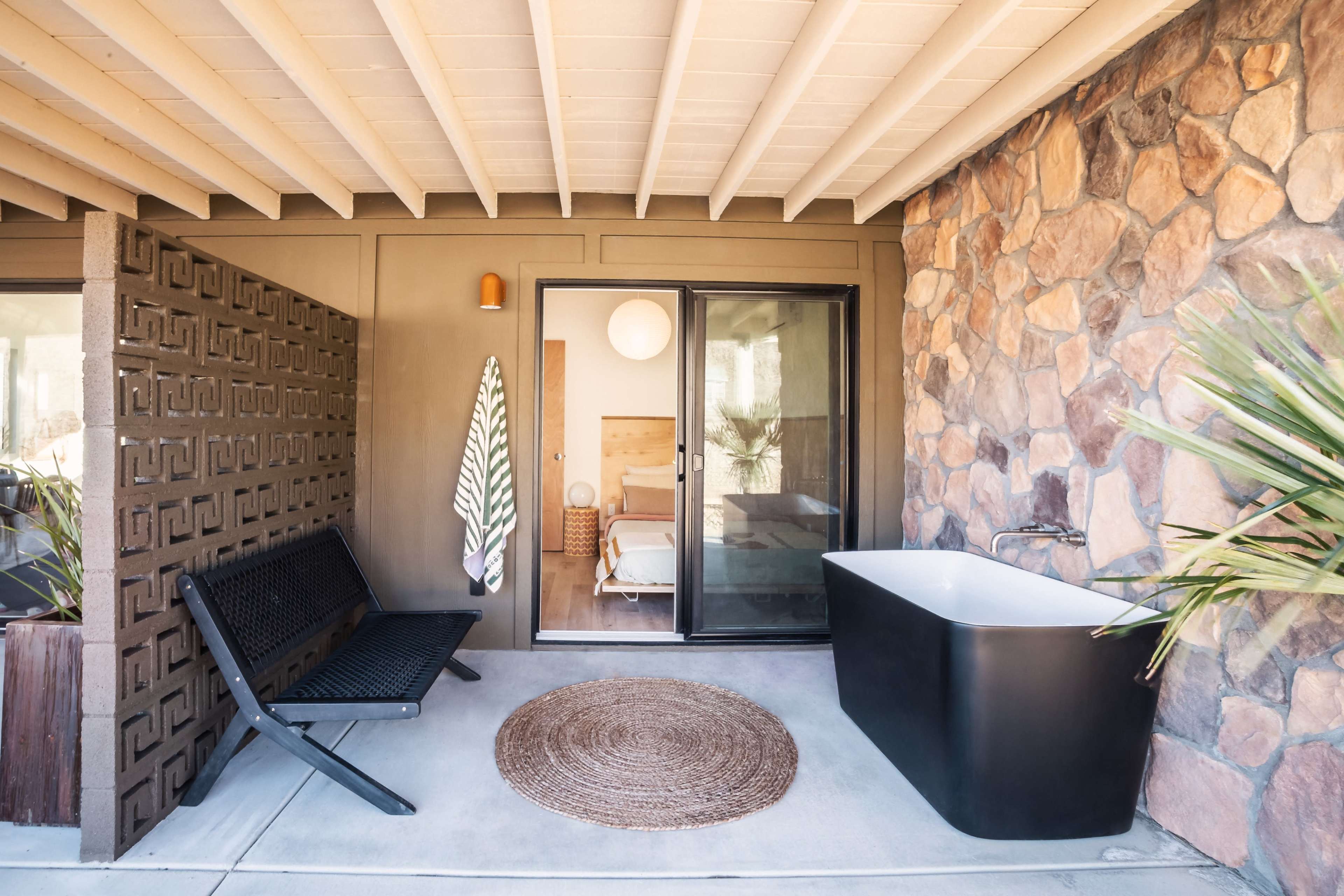A cozy outdoor area featuring a black freestanding bathtub, a woven rug, a modern bench, and a wall made of stone and decorative blocks, leading into a bedroom.