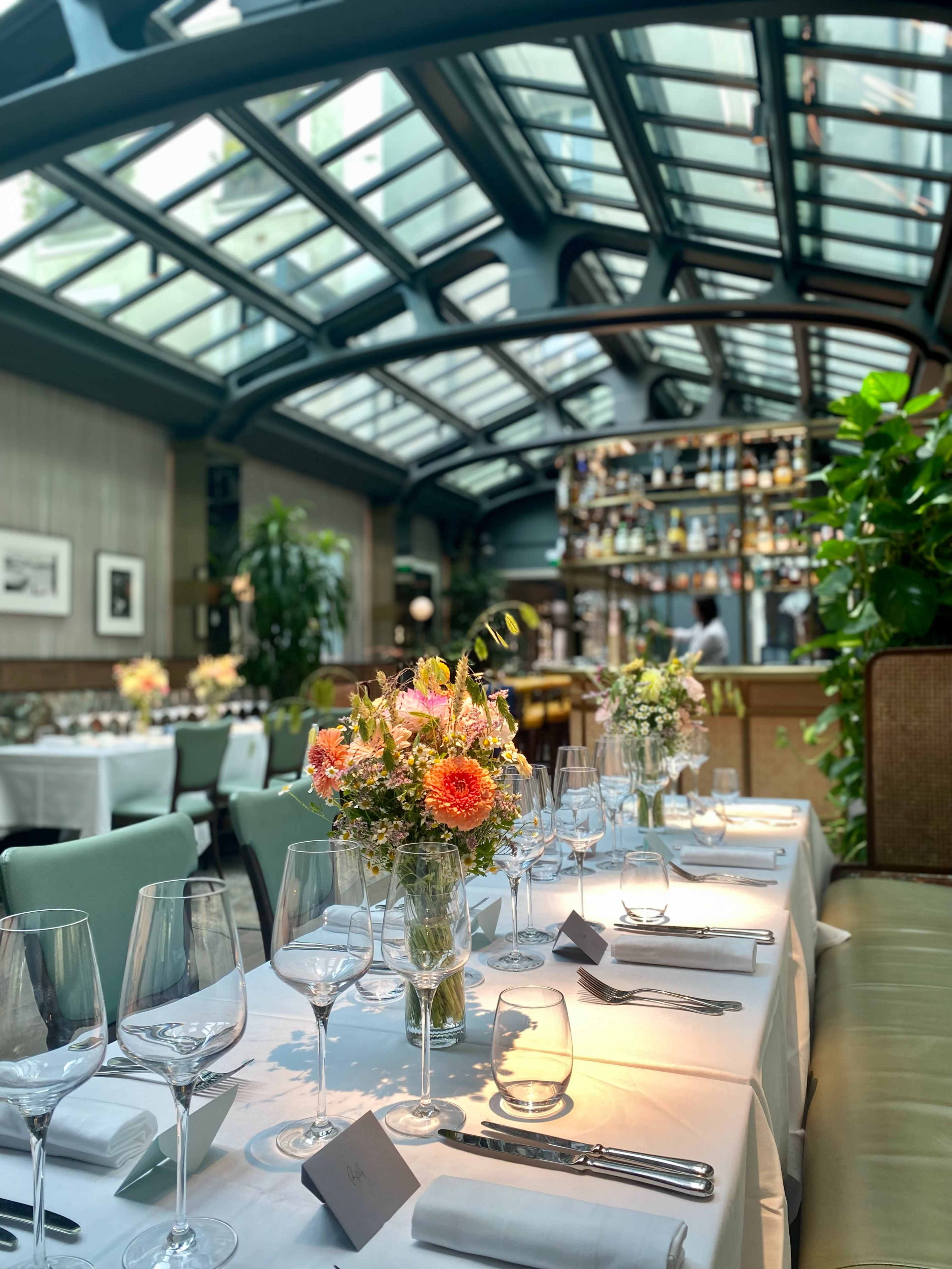 A brightly lit restaurant with a long table set for dining, featuring a vase of colorful flowers in the center and plants in the background.