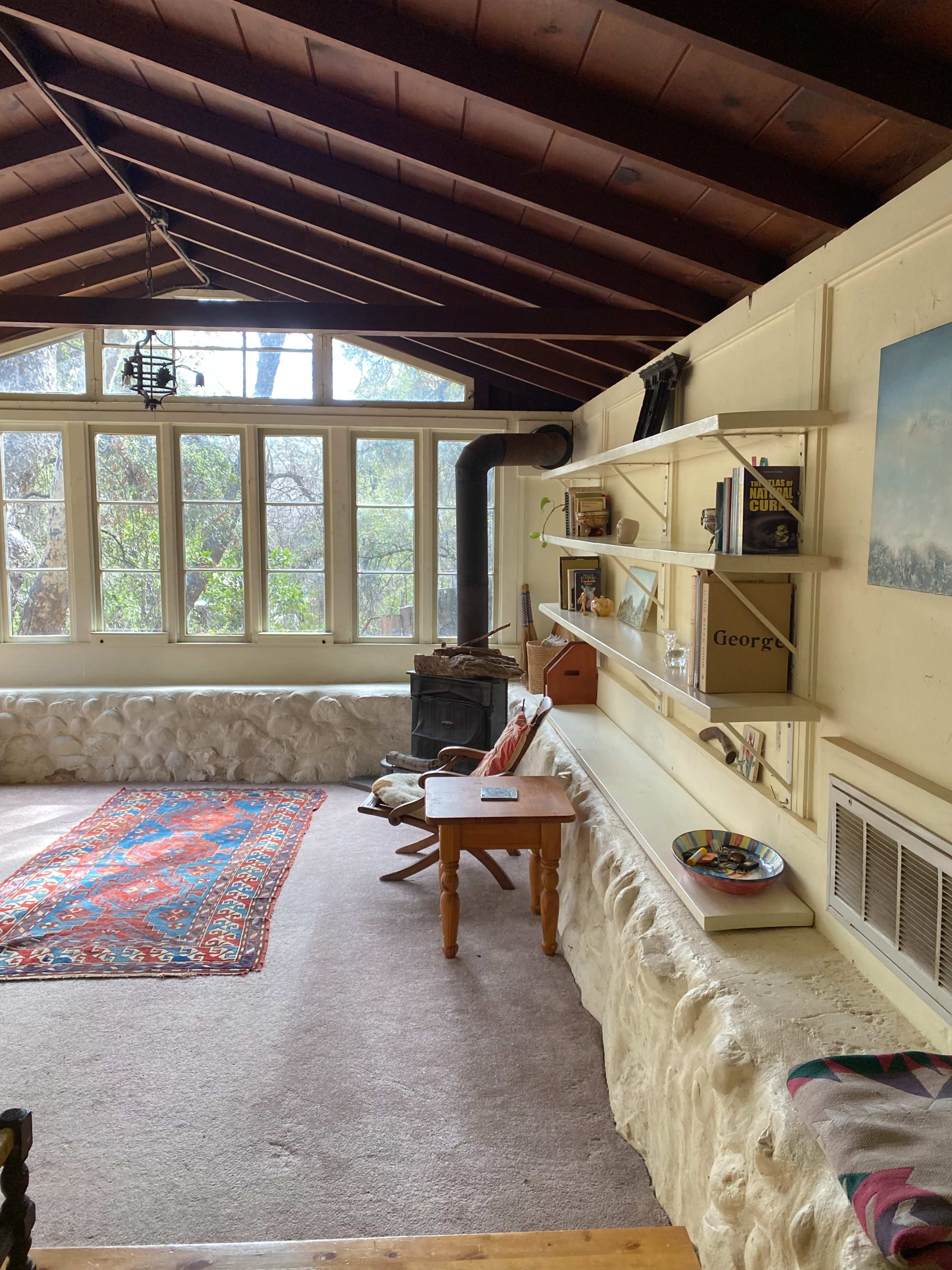 A cozy room with a stone wall, a wooden table, a bookshelf, and large windows letting in natural light.