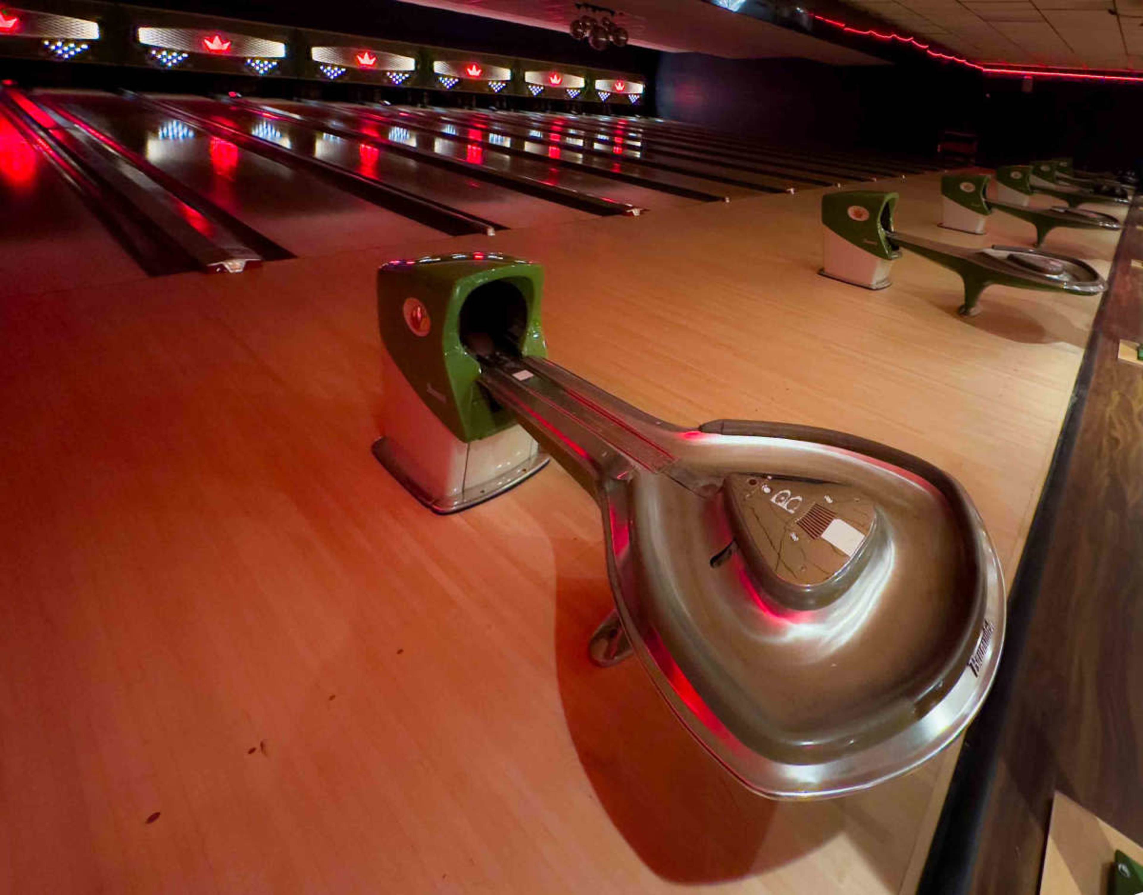 The image shows a bowling alley with a focus on a metal ball return system and several bowling lanes illuminated by overhead lights.