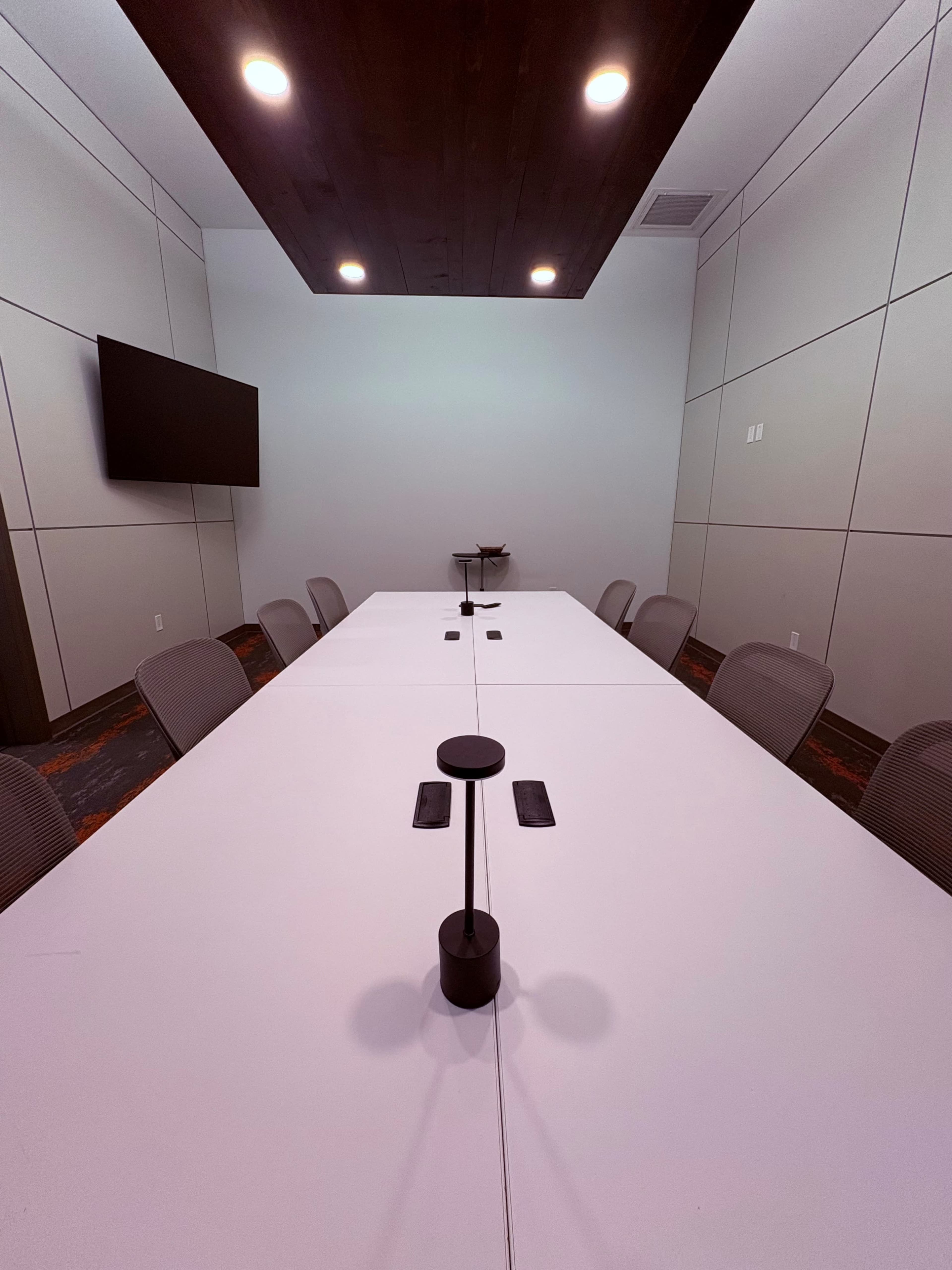 A modern conference room features a long white table surrounded by gray chairs, with a wall-mounted screen and overhead lighting.