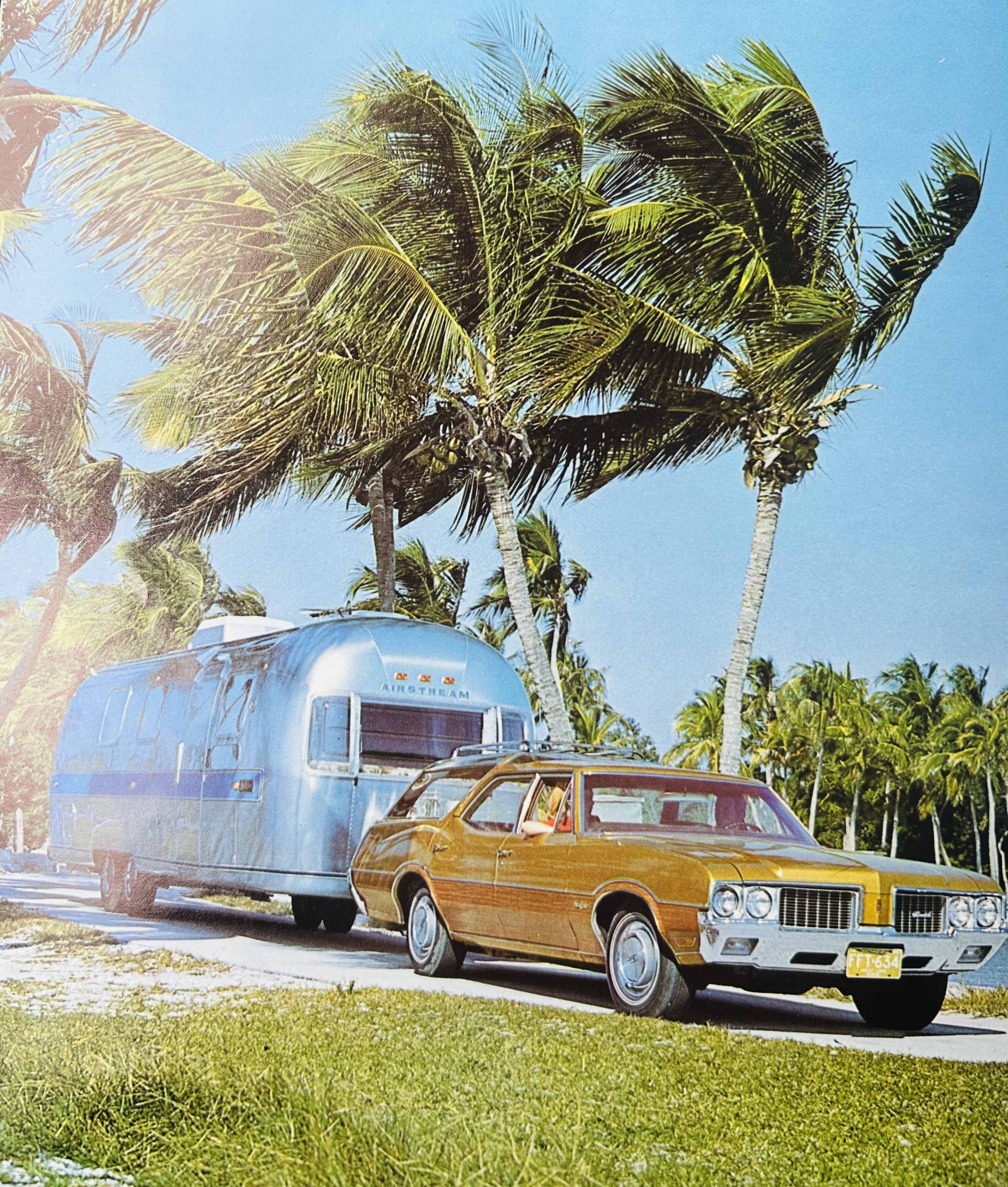 A vintage car tows a shiny trailer along a palm-lined road under a clear blue sky.