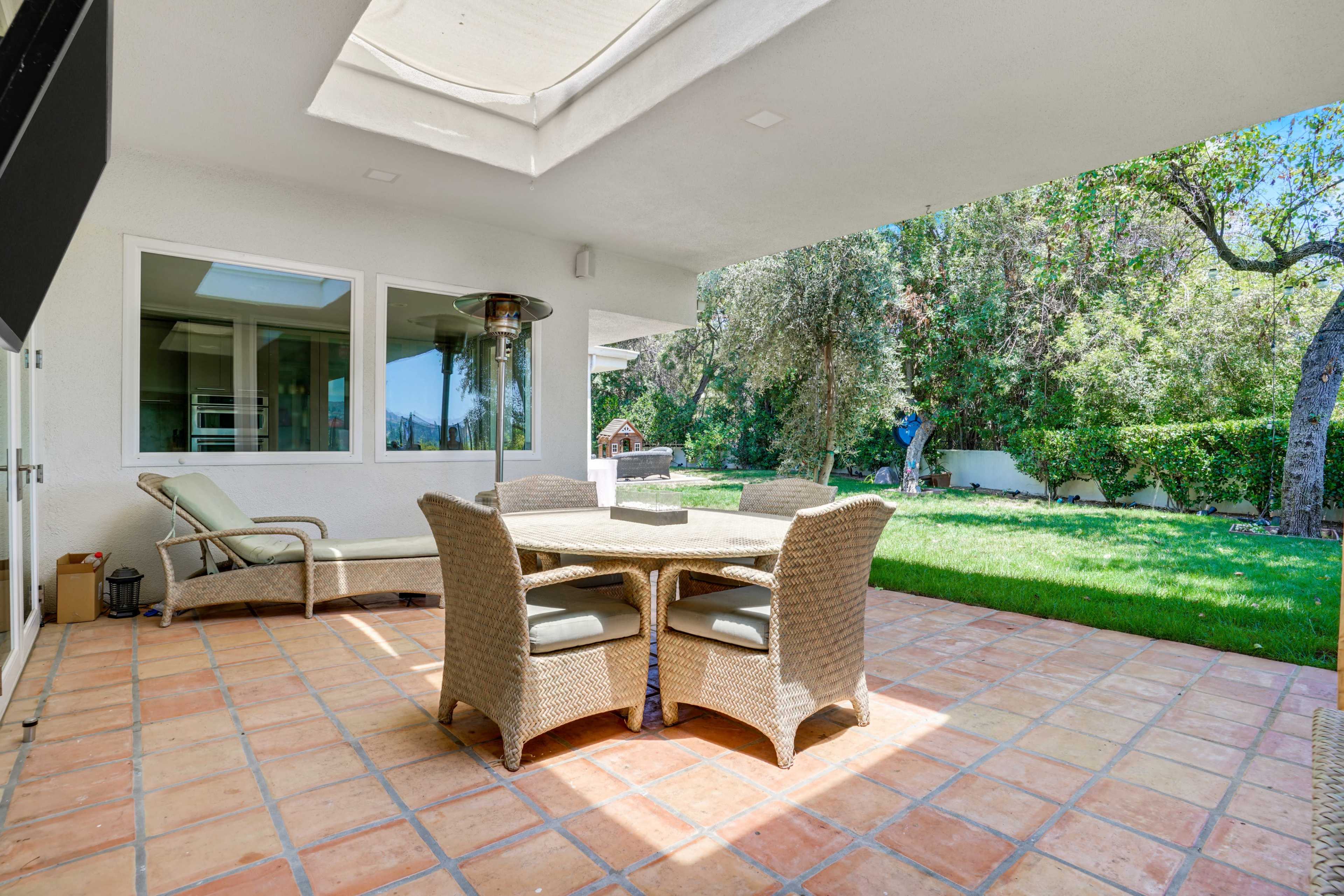 The image shows a covered patio area with a wicker dining set, surrounded by greenery and trees.