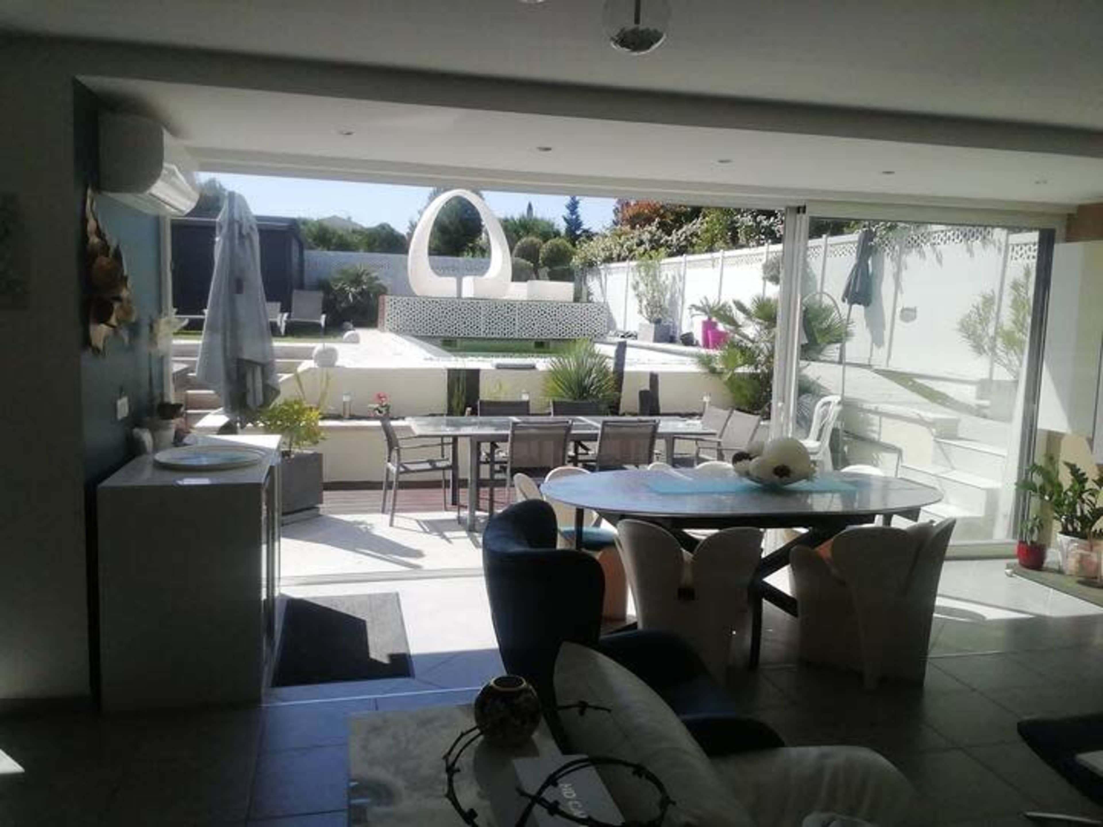 The image shows a bright interior space with large glass doors opening to an outdoor area featuring a pool, modern sculptures, and various plants.