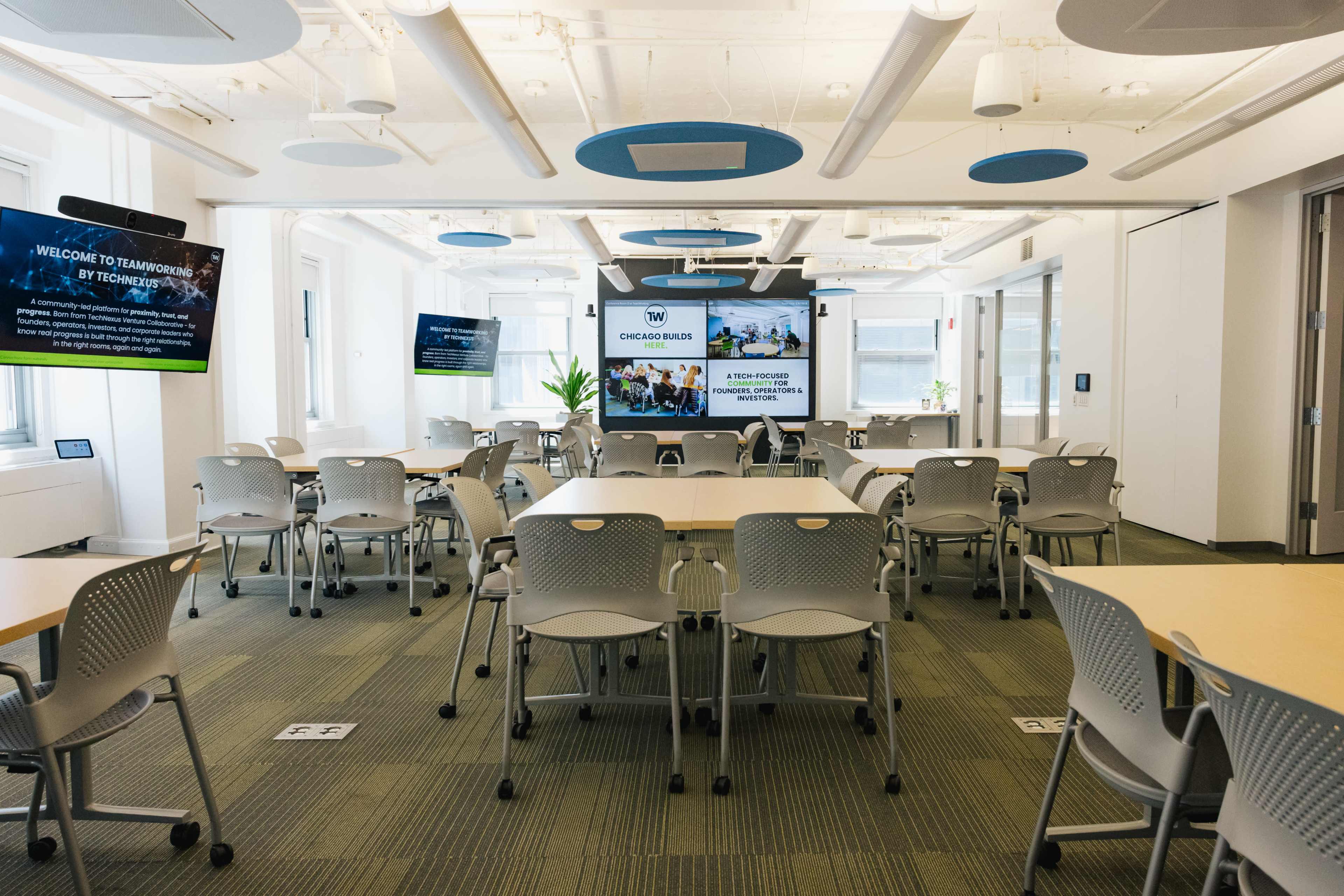 A spacious conference room is set up with multiple tables and chairs, featuring large screens displaying welcoming messages and visuals.