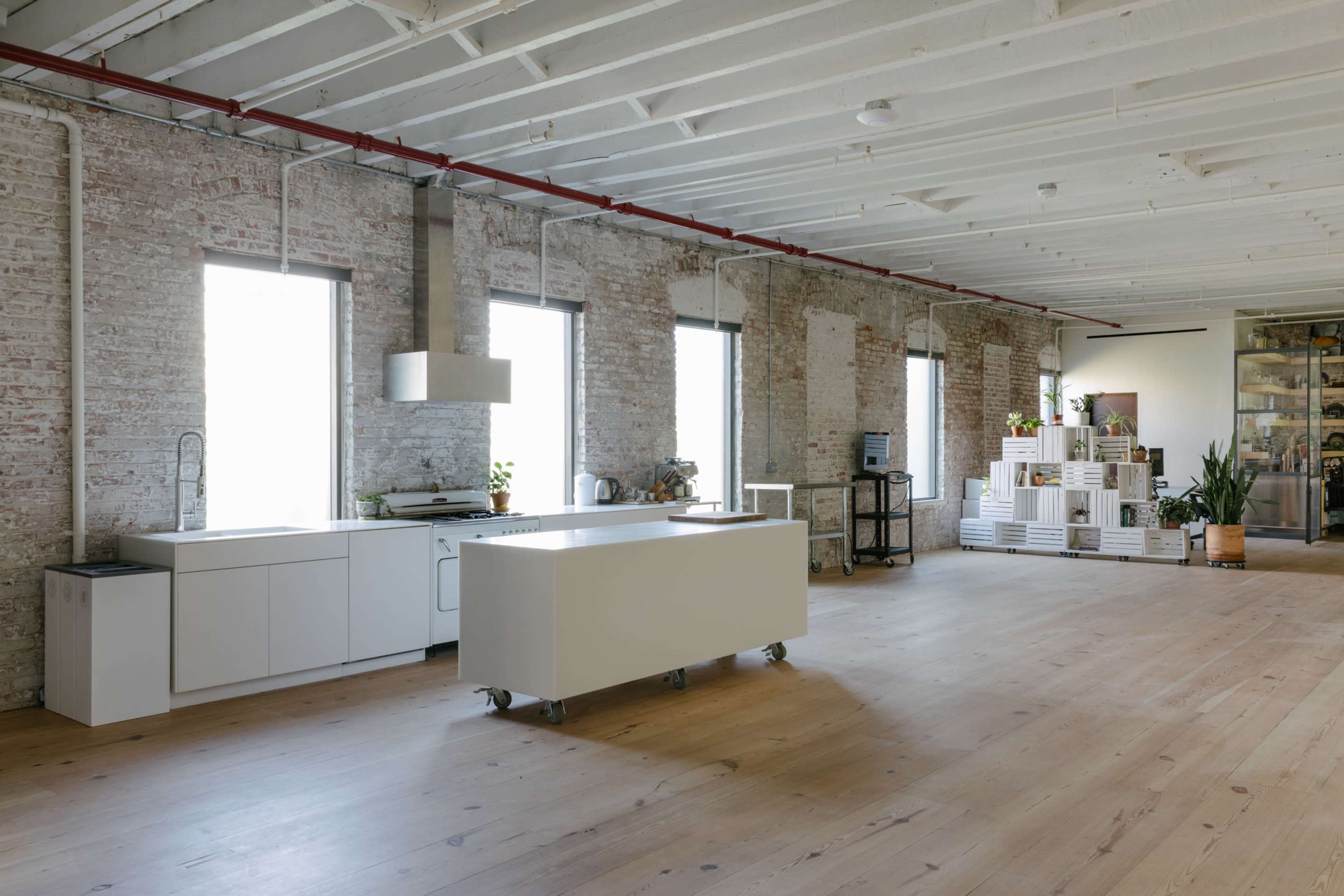 The image shows a spacious industrial-style kitchen area with white brick walls, large windows, and a central island on wheels.