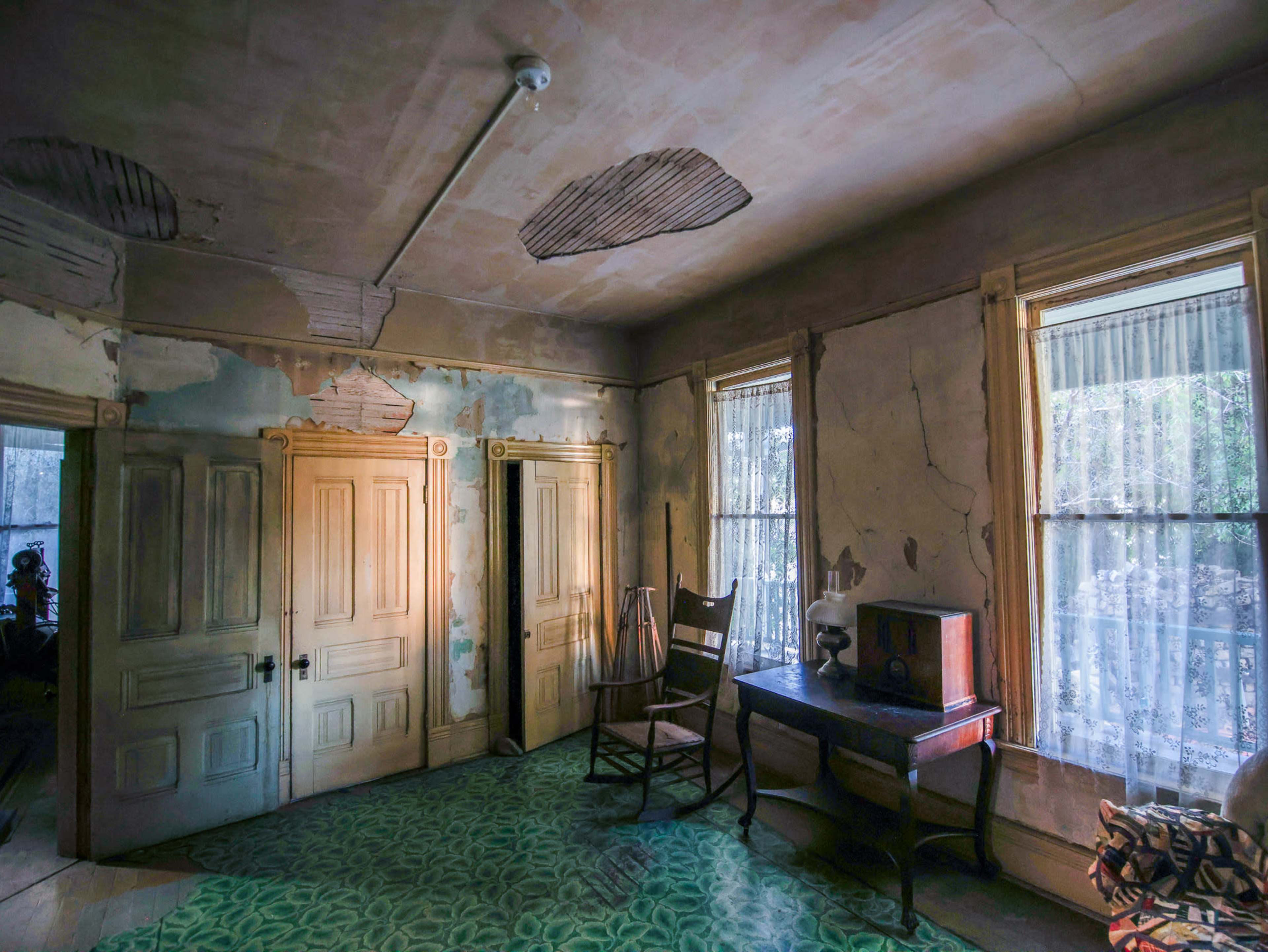 The image shows a dilapidated room with peeling walls, a wooden chair, a desk, and two windows adorned with curtains.