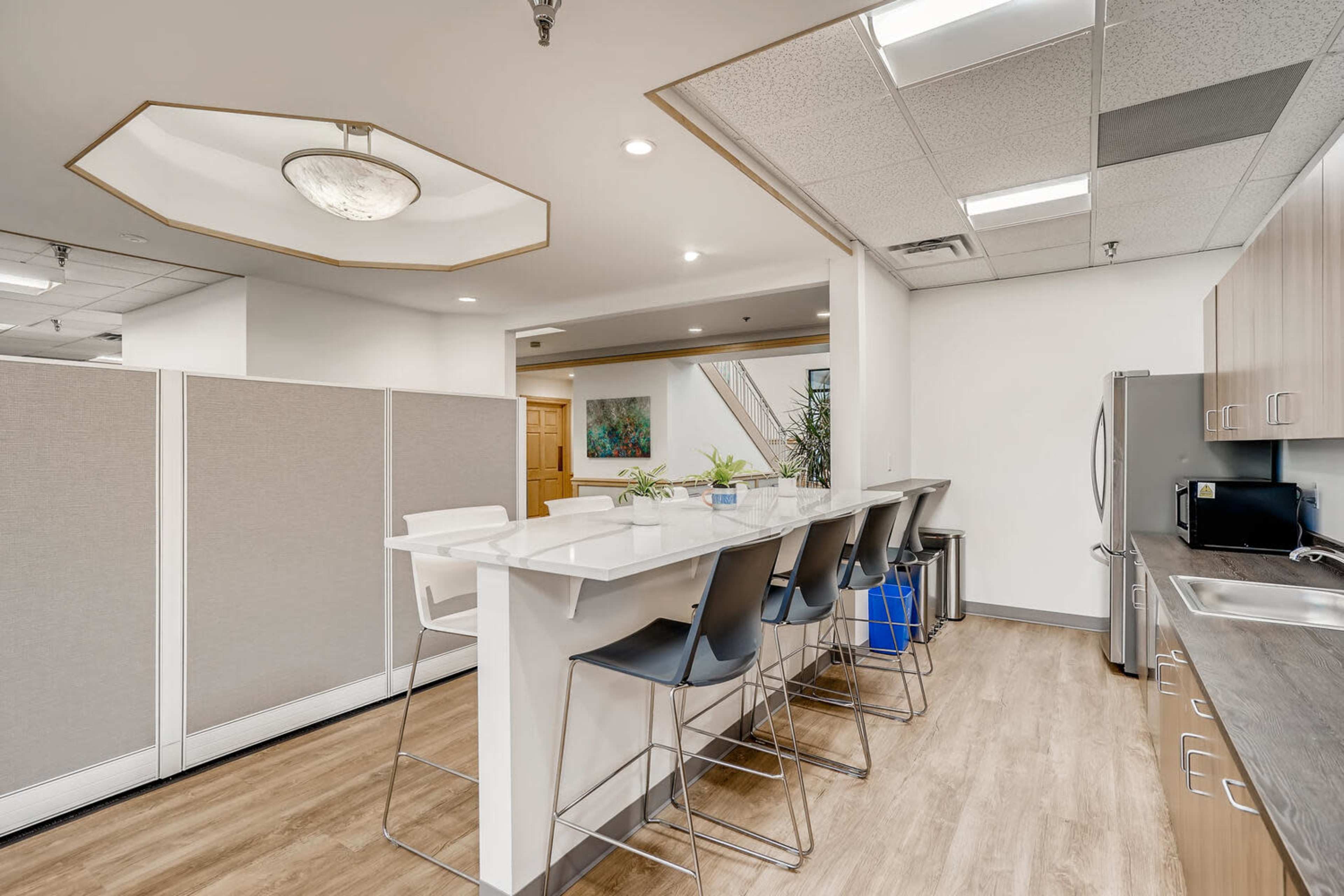 A modern kitchenette features a white countertop with high stools, stainless steel appliances, and partitioned walls.