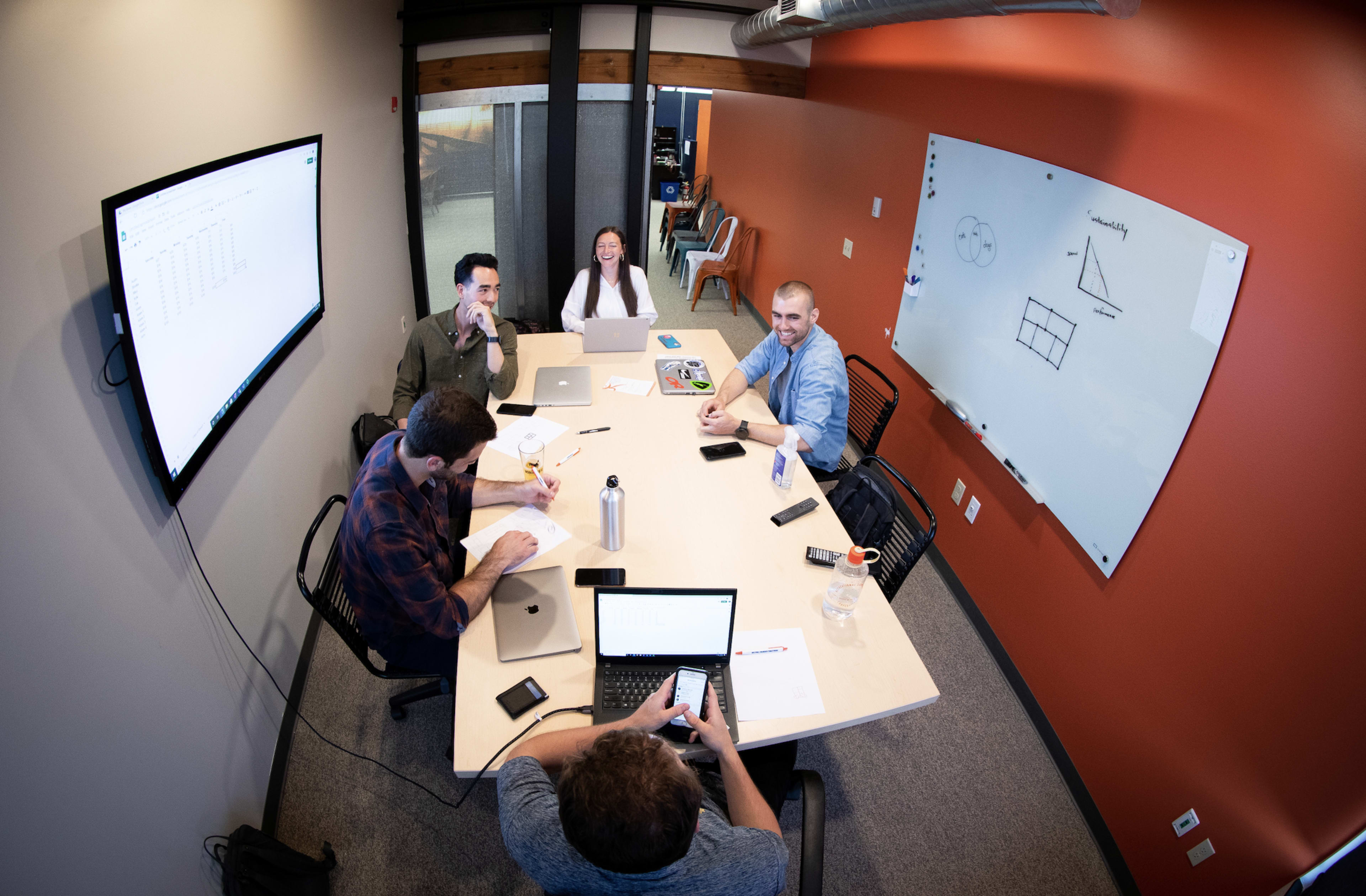 A group of five people is sitting around a conference table with laptops and documents, engaged in a meeting in a modern office setting.