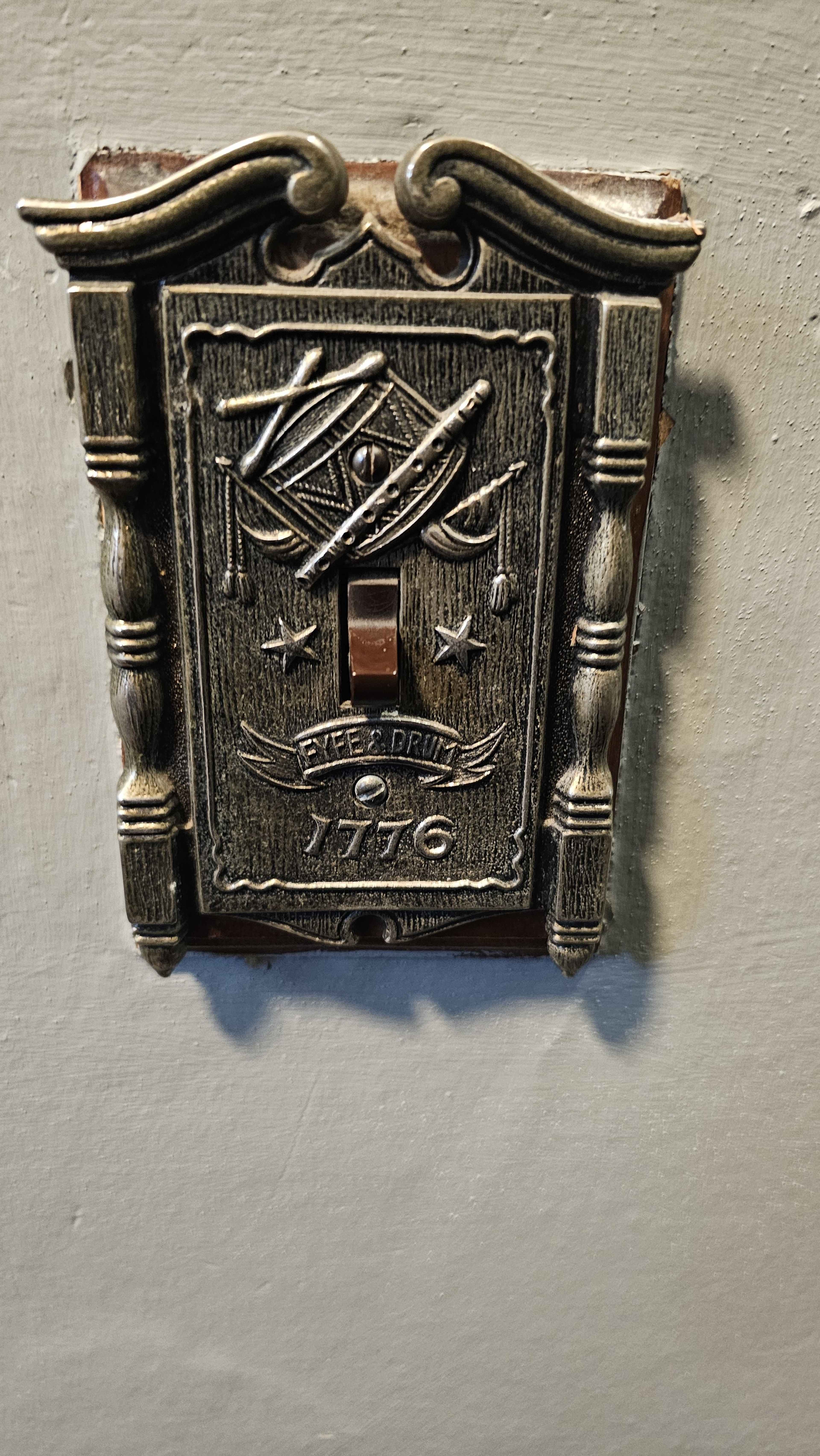 The image shows a decorative light switch cover featuring Masonic symbols, set against a plain wall.
