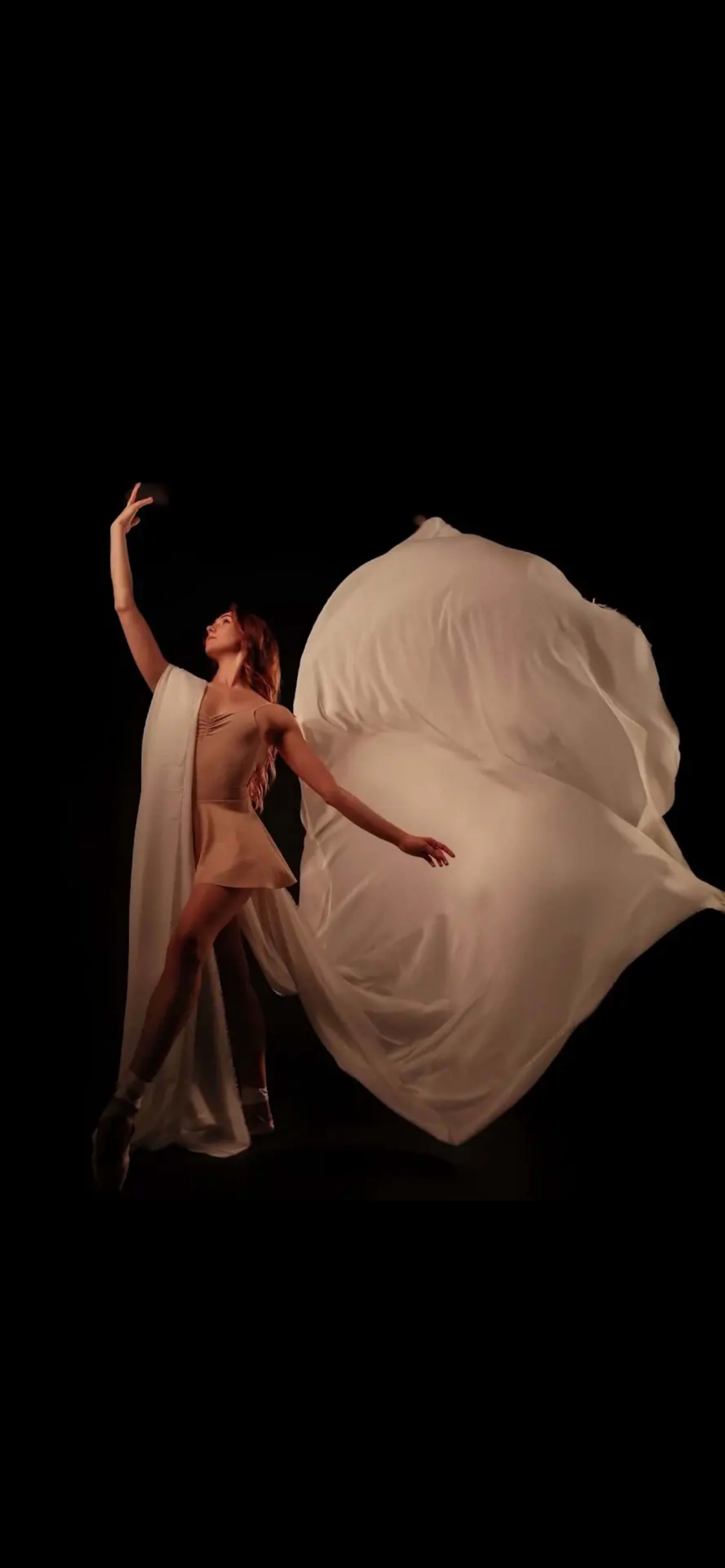 A dancer performs gracefully in a dramatic pose, wearing a skin-toned outfit and flowing white fabric against a dark background.