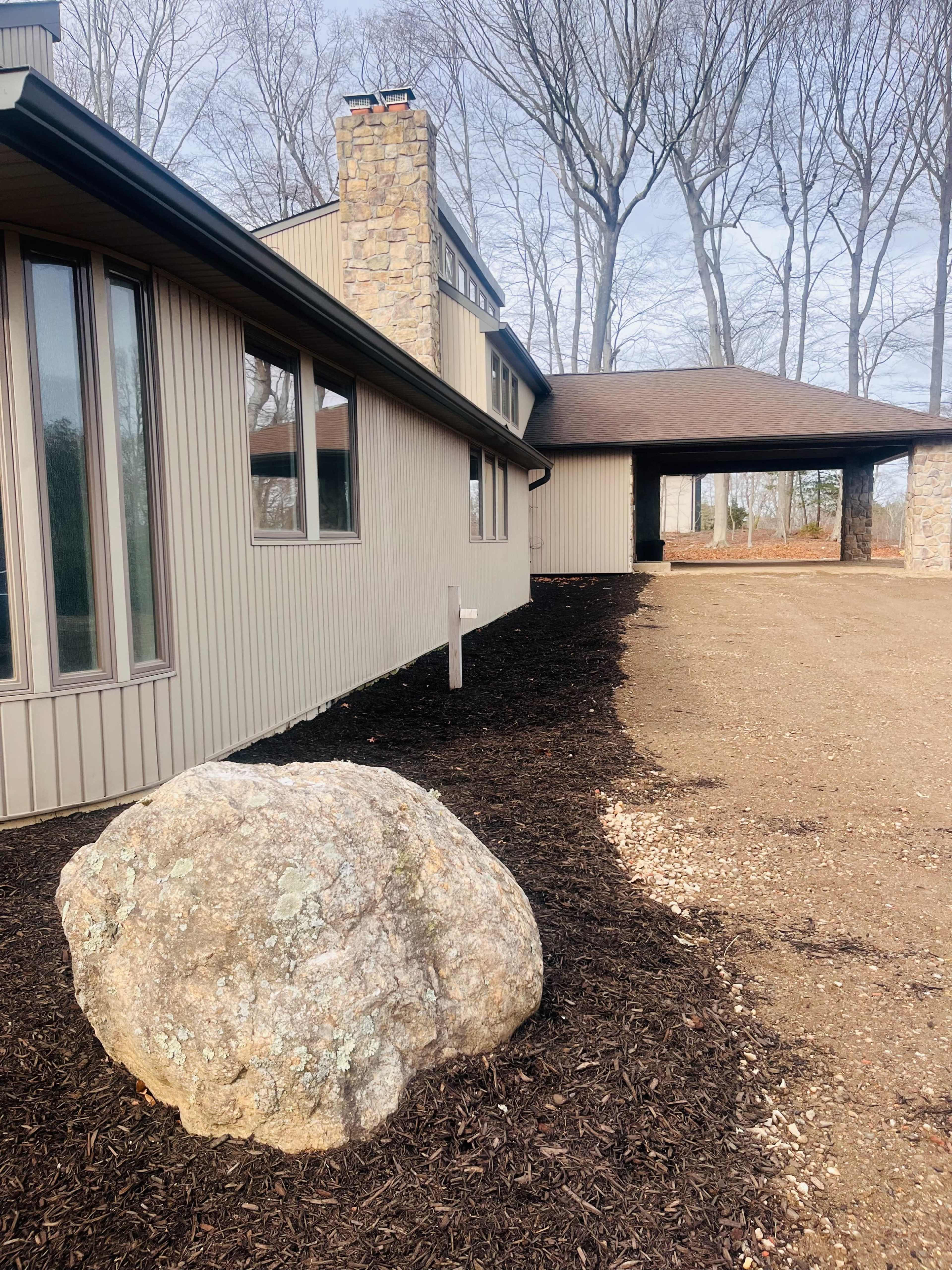A large boulder sits in front of a modern house with a stone chimney, surrounded by freshly mulched landscaping.