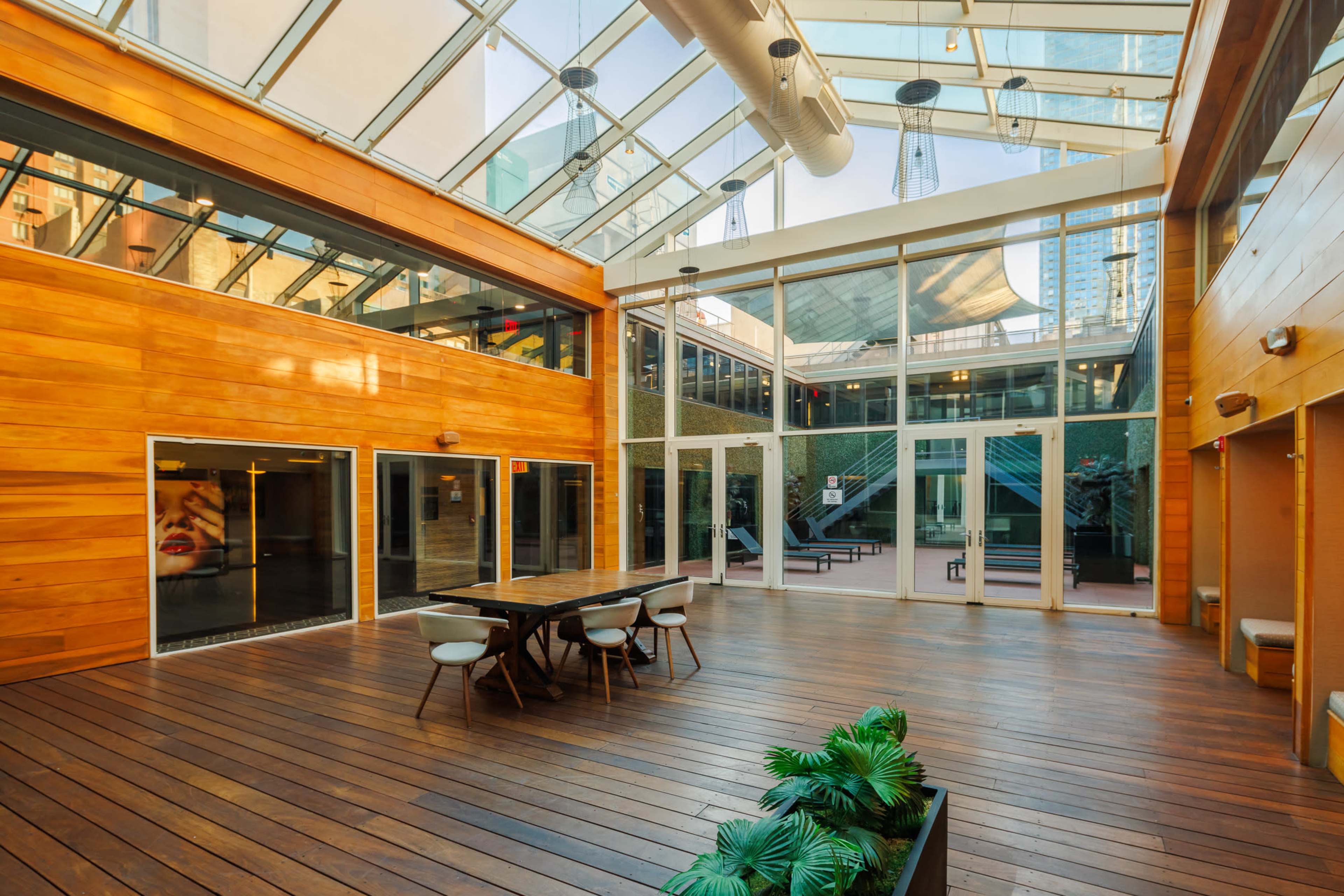 The image shows a spacious indoor area with wooden walls and a glass ceiling, featuring a table and chairs surrounded by greenery.