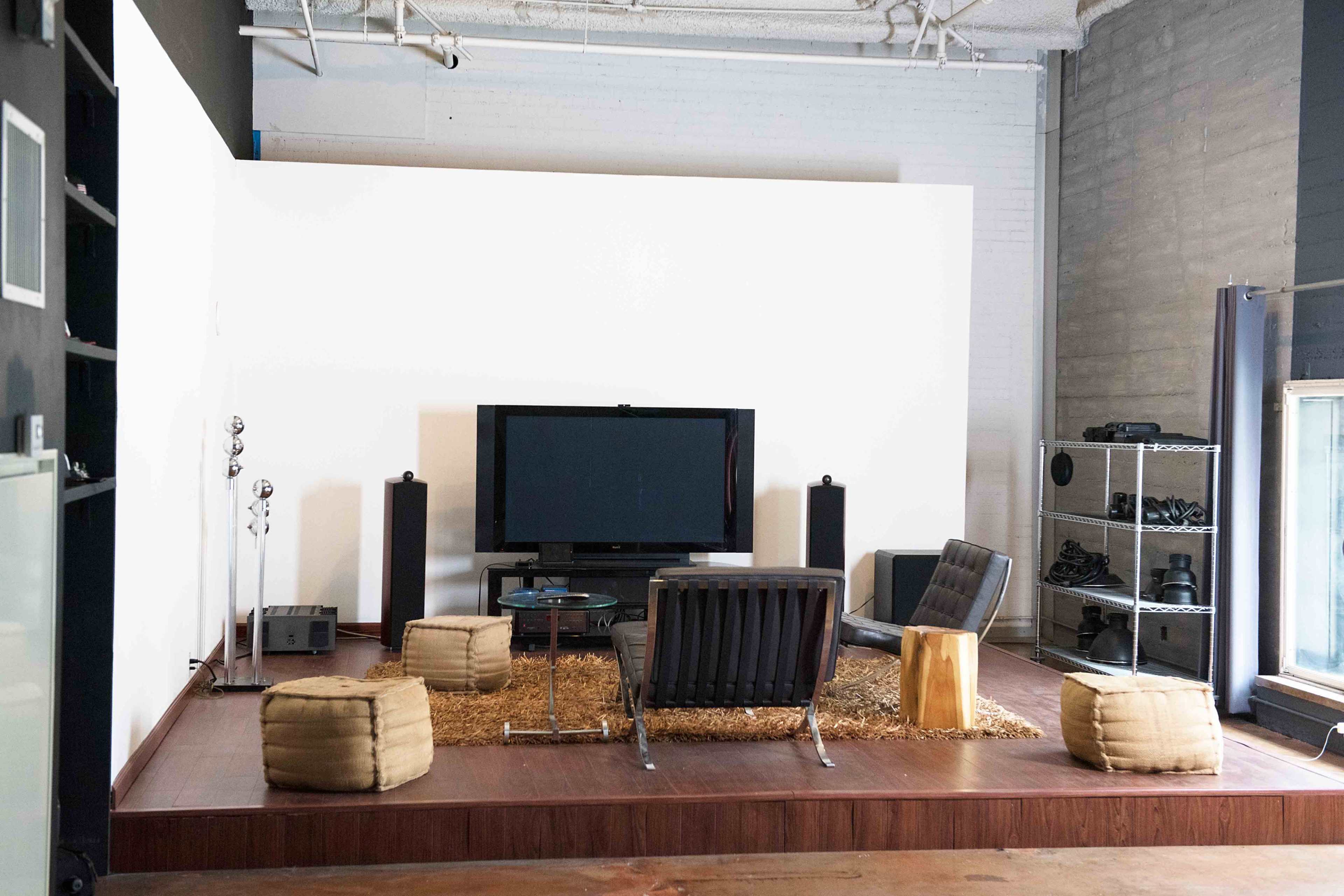 A modern living room setup features a large television on a white wall, surrounded by minimalist furniture, including two black chairs, a coffee table, and several seating poufs on a wooden floor.