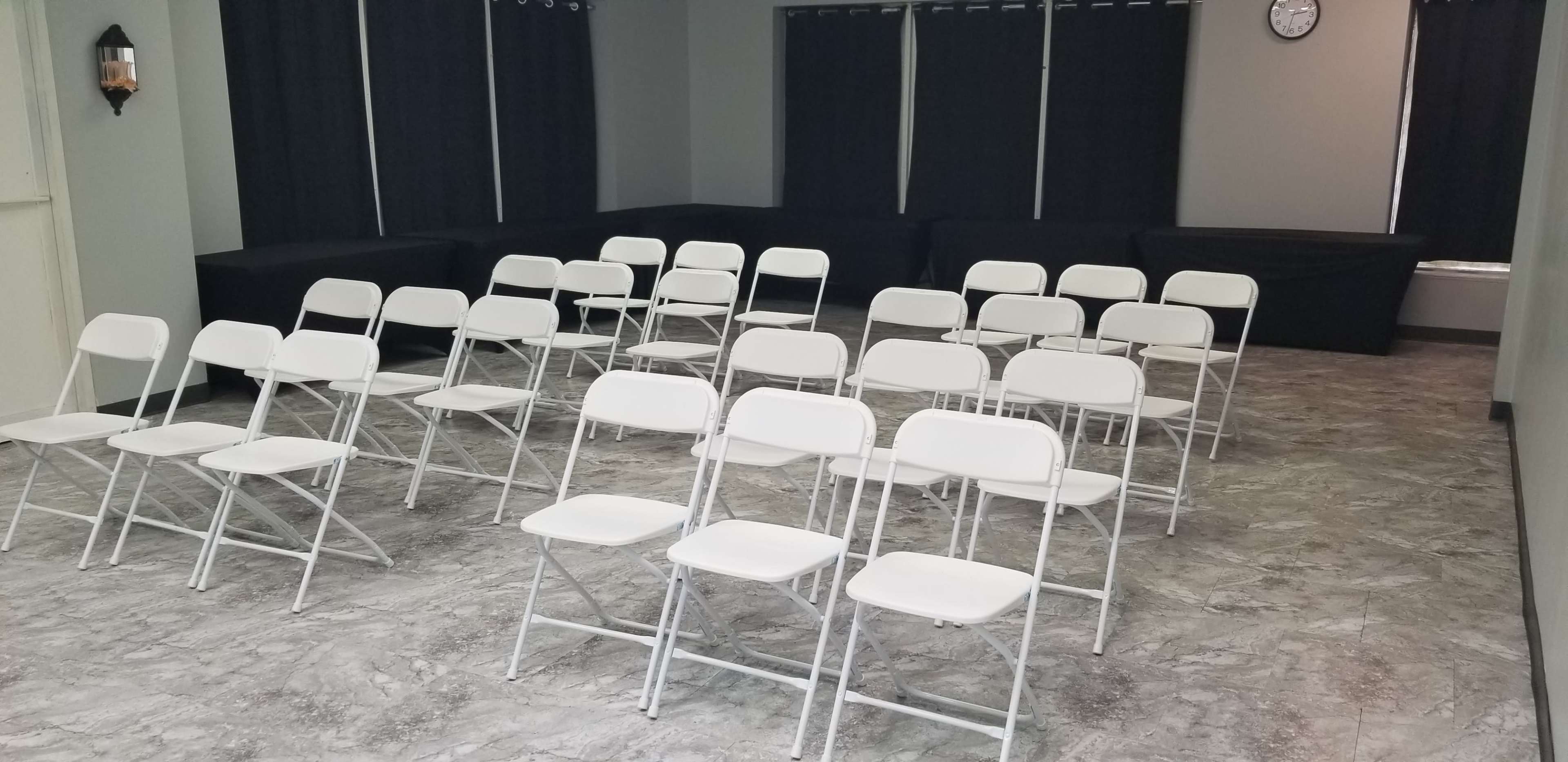 The image shows a room with gray flooring and black curtains, arranged with rows of white folding chairs.