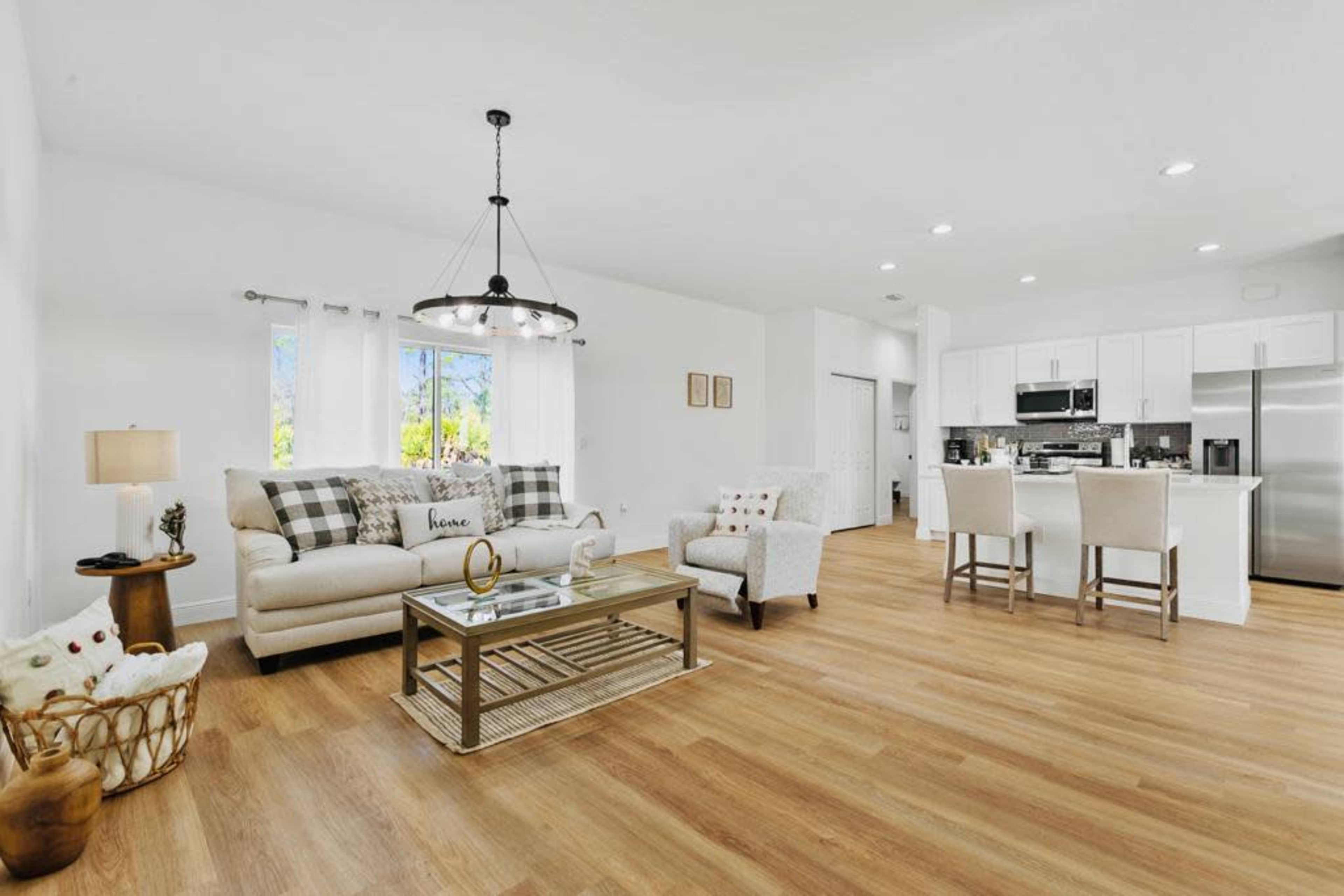 The image shows a modern living room with a light-colored sofa, a glass coffee table, and an open kitchen area featuring white cabinets and stainless steel appliances.