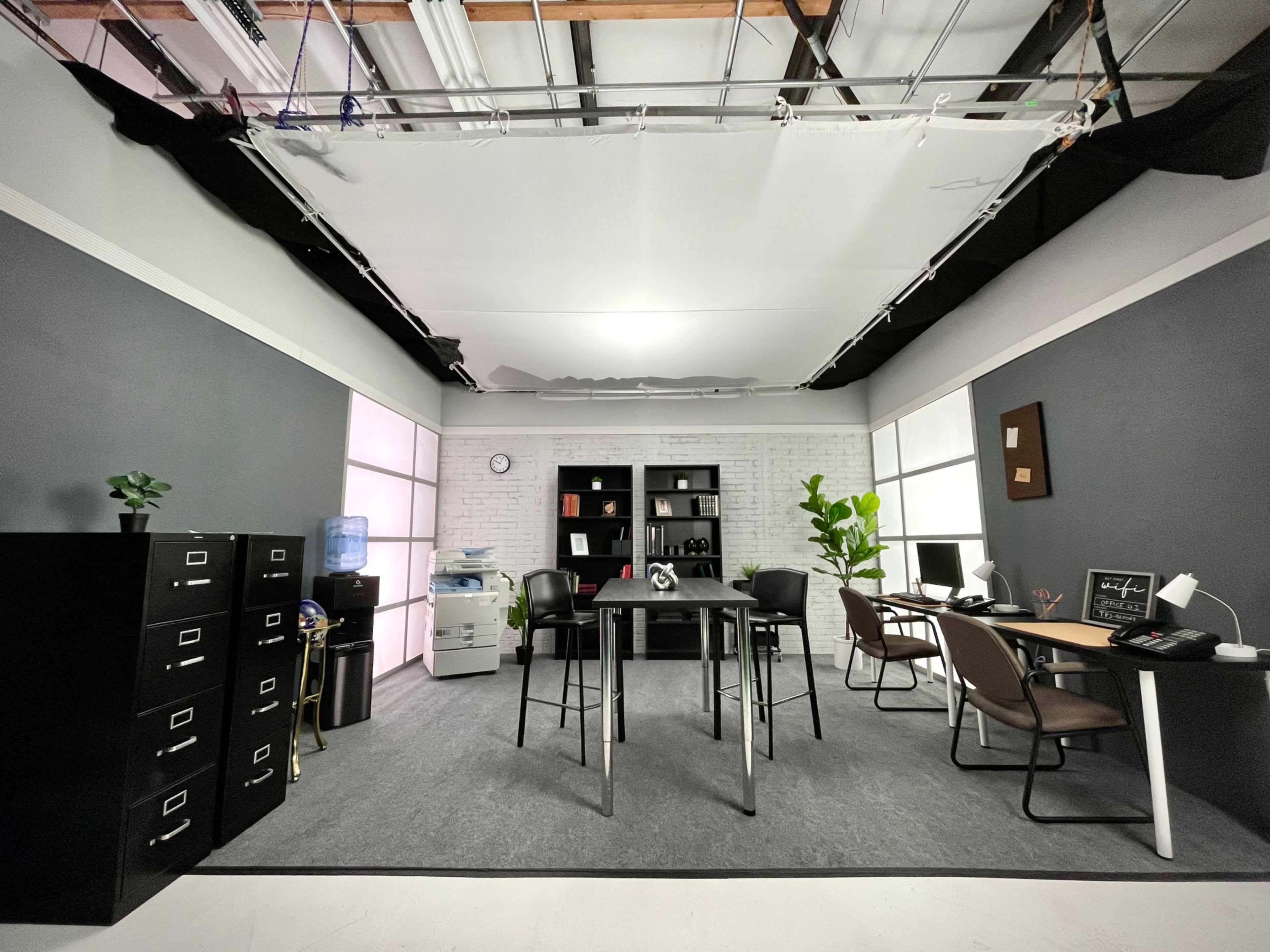 A modern office space with a central table, filing cabinets, chairs, and a desk setup against a backdrop of gray walls and large windows.