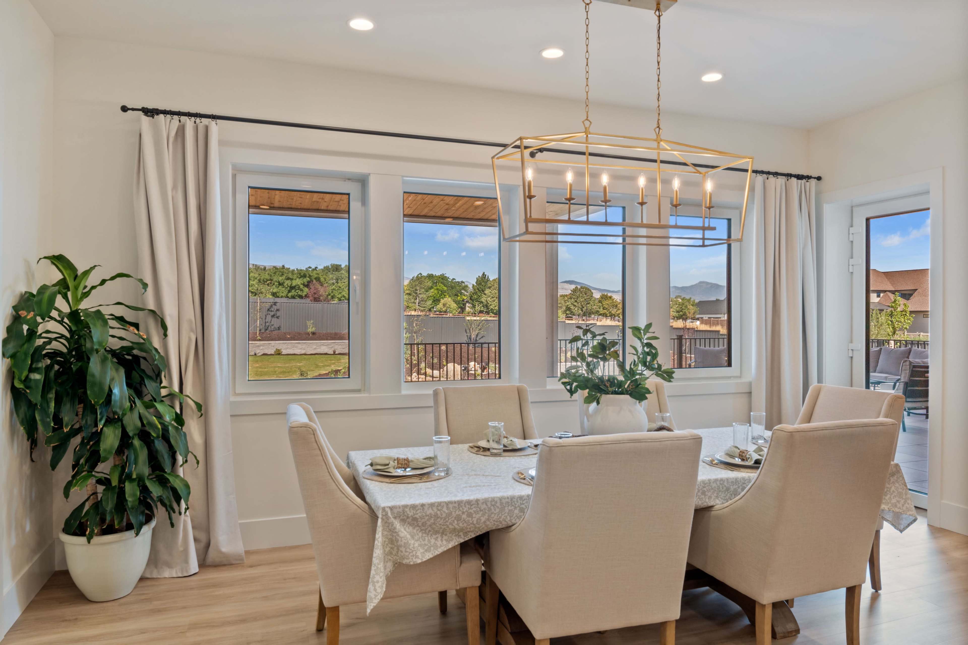 A light-filled dining area features a large table set for a meal, surrounded by beige upholstered chairs and a modern light fixture overhead, with views of a scenic outdoor landscape through large windows.