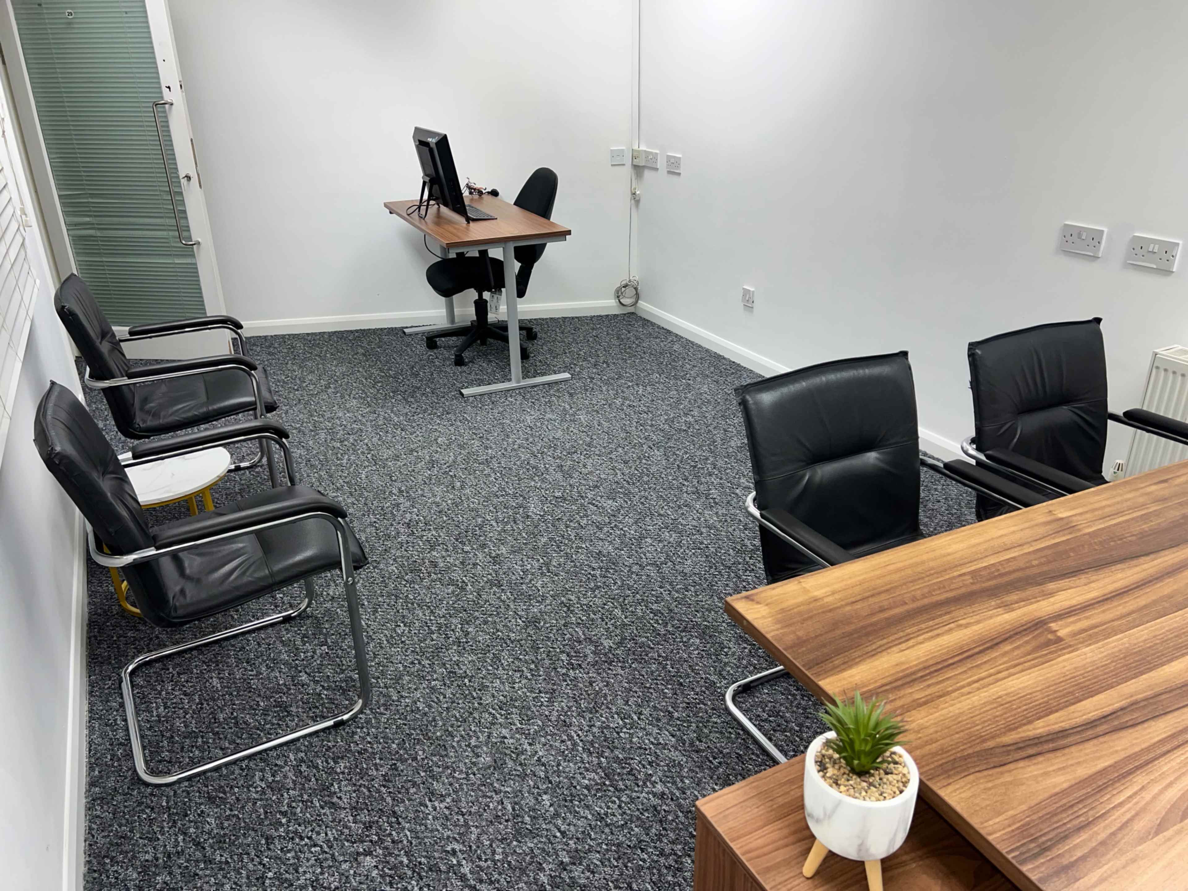 The image shows a small, empty office space featuring a desk with two monitors, several black chairs, and a potted plant on a wooden table.