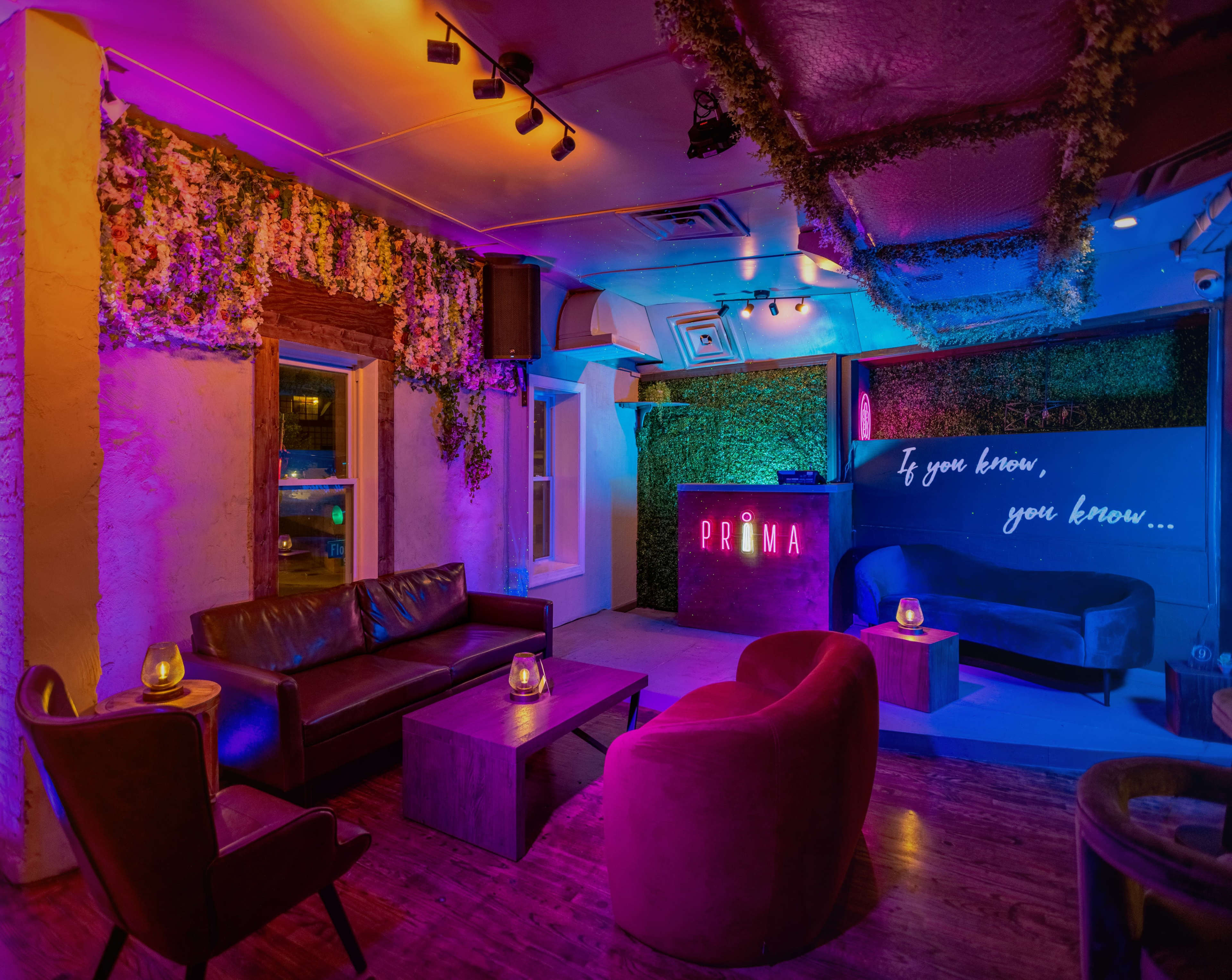 A dimly lit lounge with colorful lighting, featuring plush seating arrangements and decorative floral elements on the walls.
