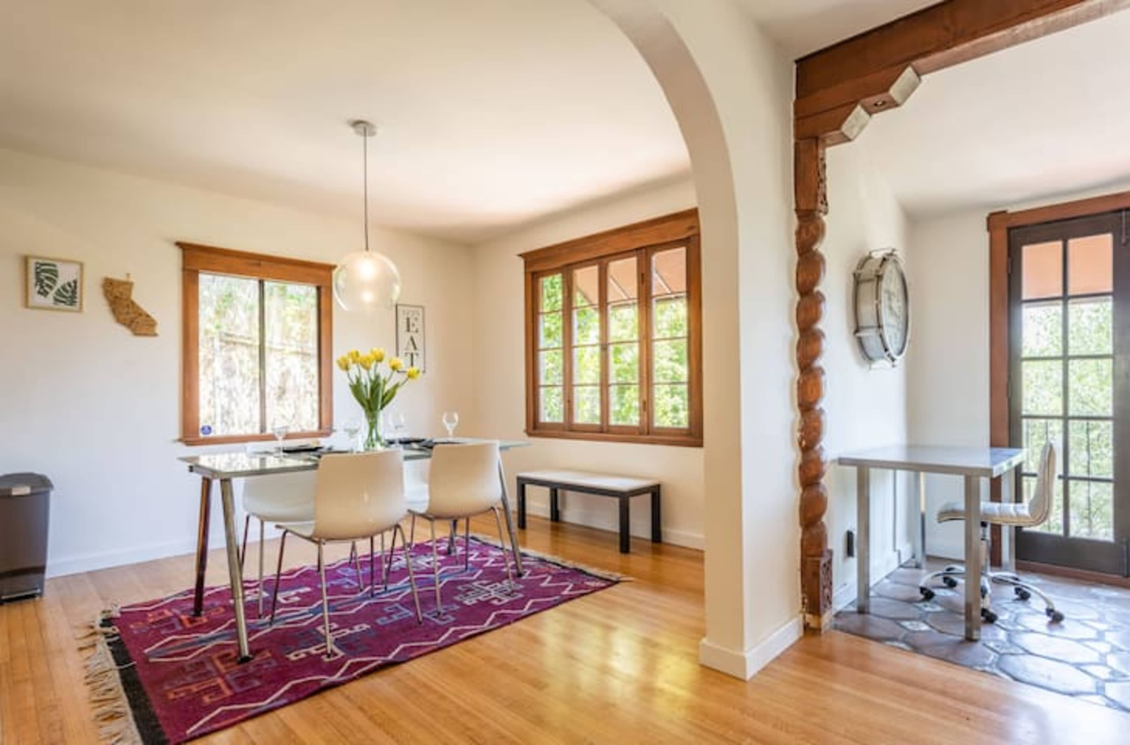 A bright dining area features a glass table surrounded by white chairs, with large windows providing natural light and a view of greenery.