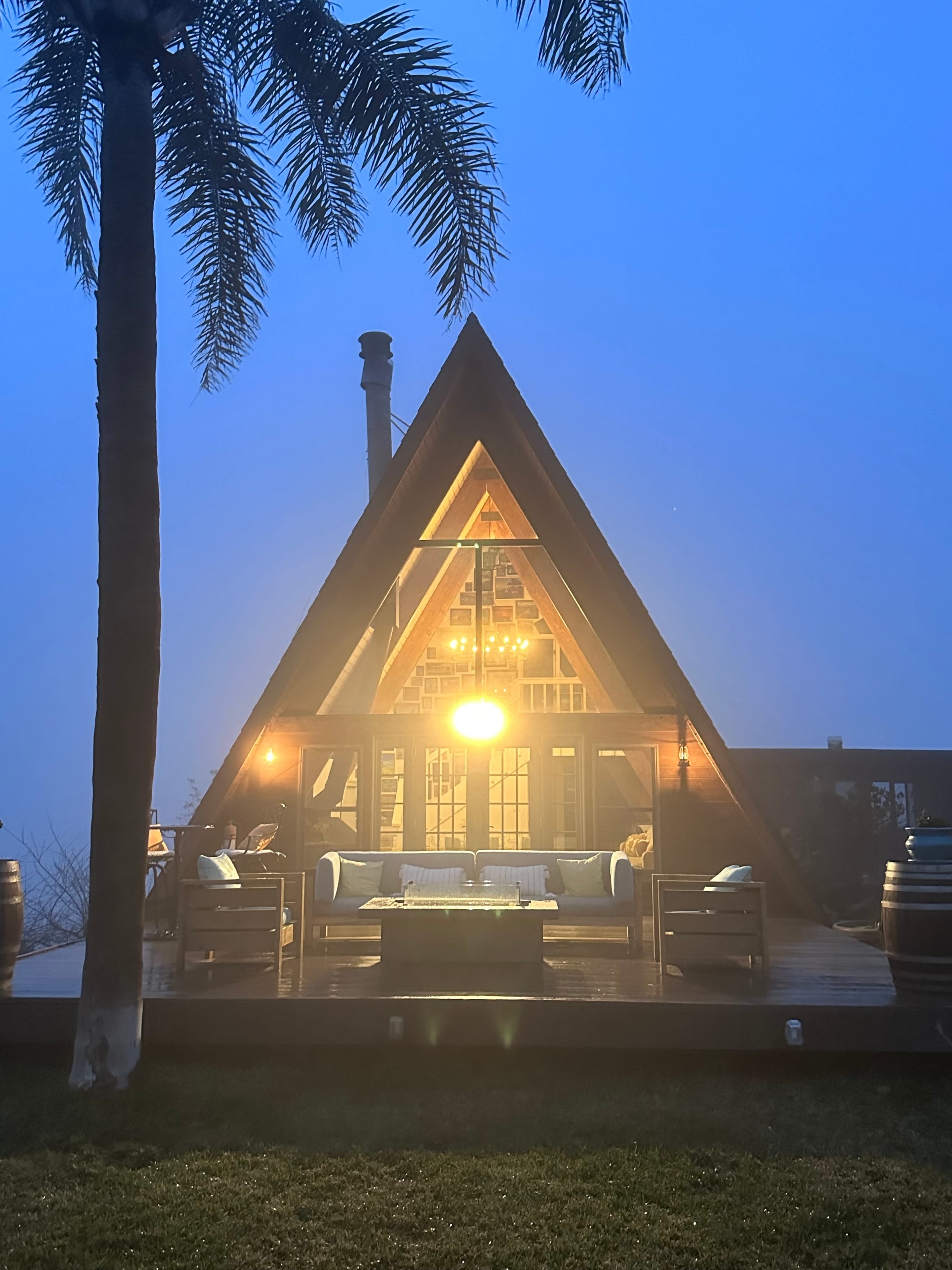 A foggy evening scene features a triangular-roofed cabin with illuminated windows and outdoor seating, surrounded by palm trees.