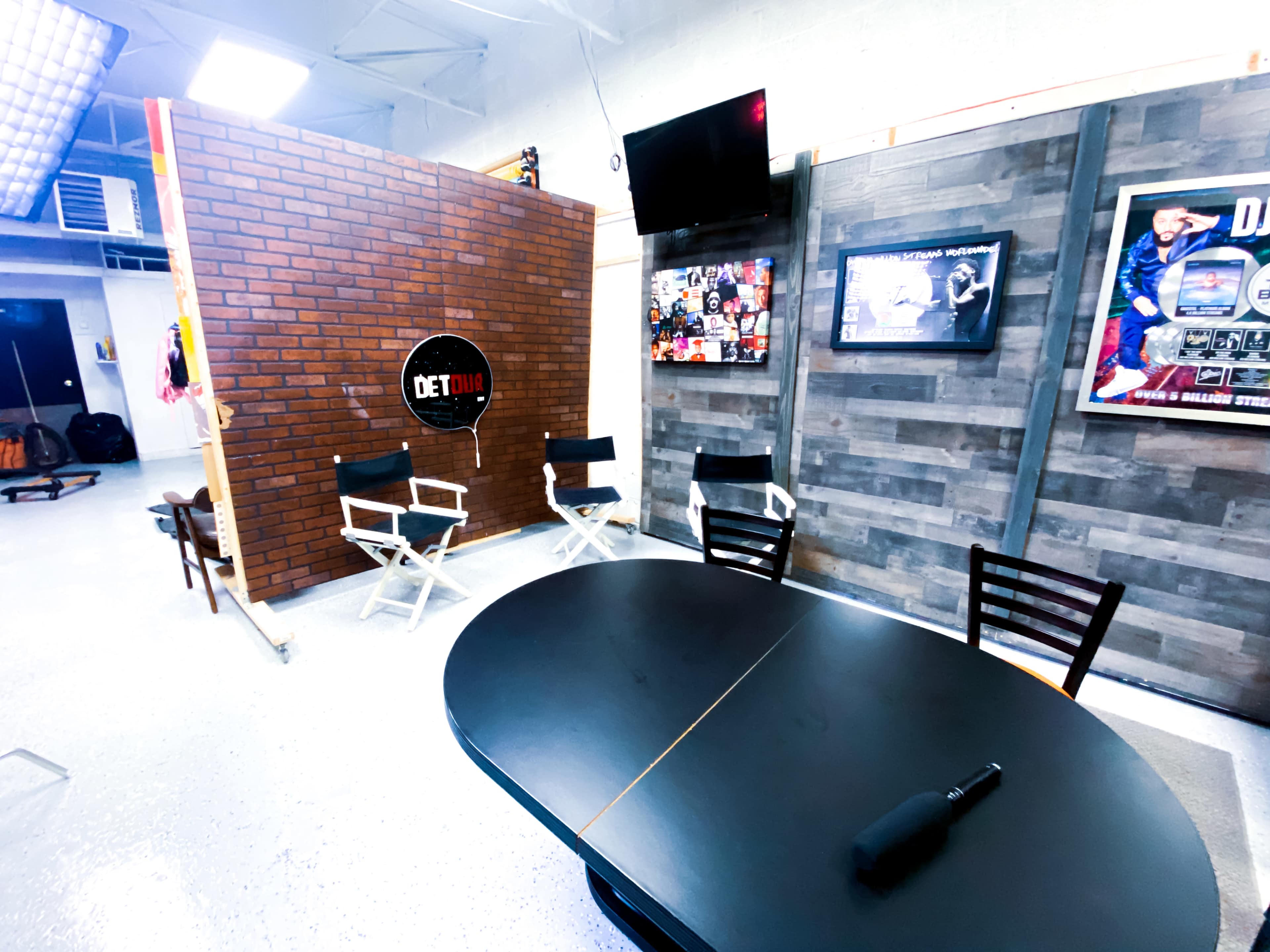 The image shows a studio space with a brick wall backdrop, a black circular table, director's chairs, and a television displaying photos and graphics on the walls.