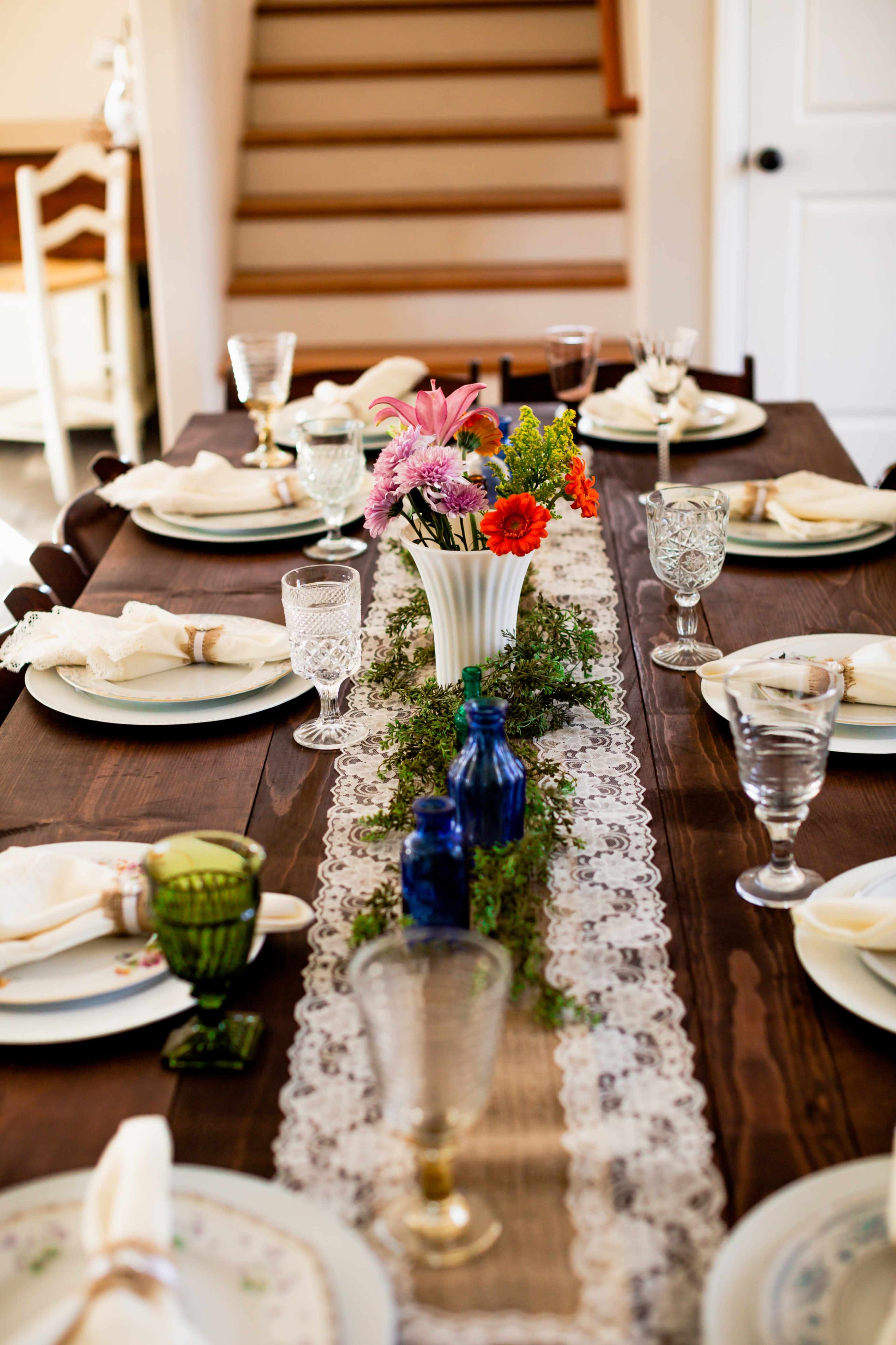 A dining table is set for a meal with white plates, glassware, and a vase of flowers on a lace table runner, with a staircase in the background.
