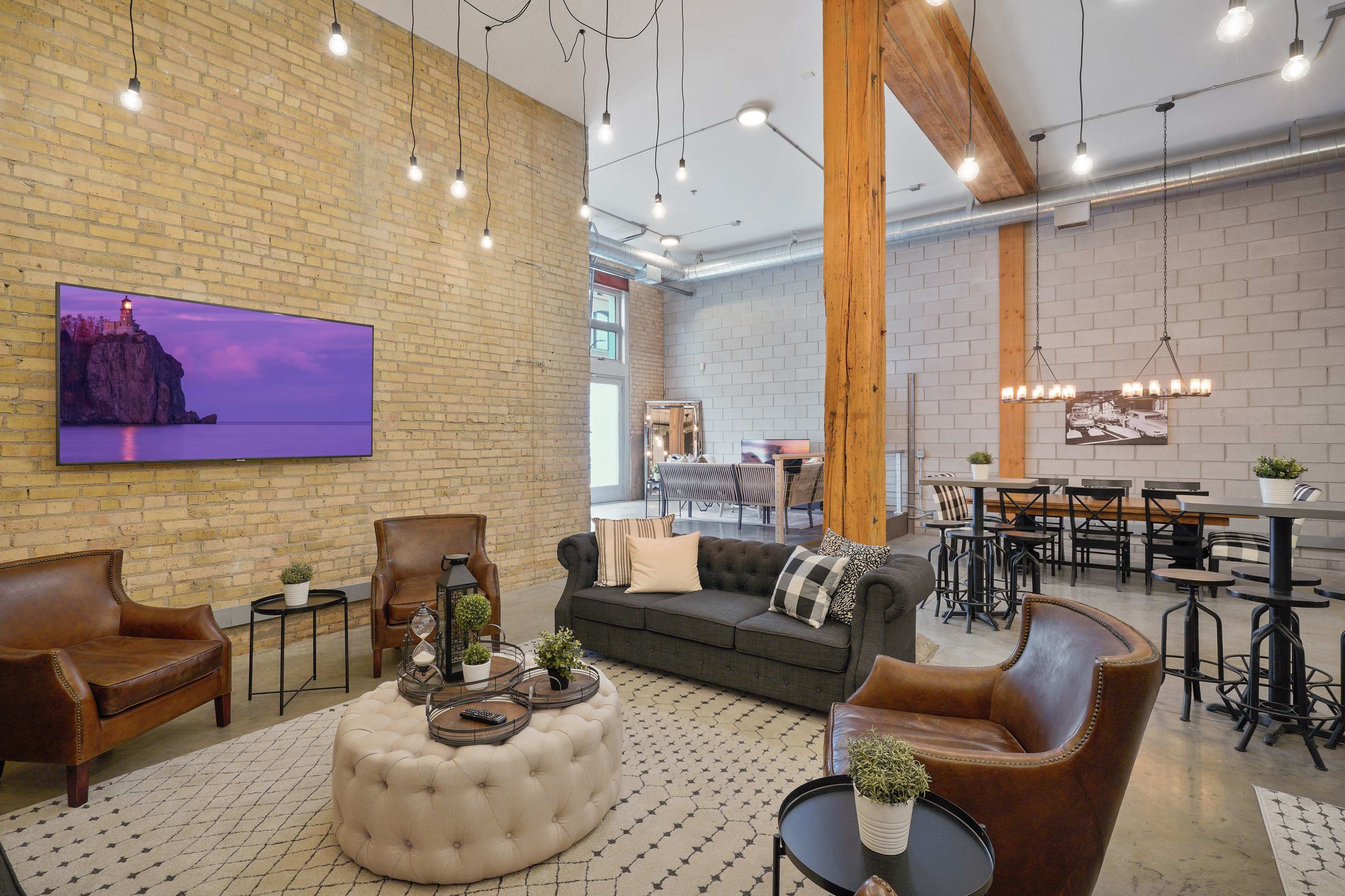 A modern industrial living space featuring a dark sectional sofa, leather armchairs, a round coffee table, and a mounted television against a brick wall, illuminated by pendant lights and surrounded by a dining area with high tables and chairs.