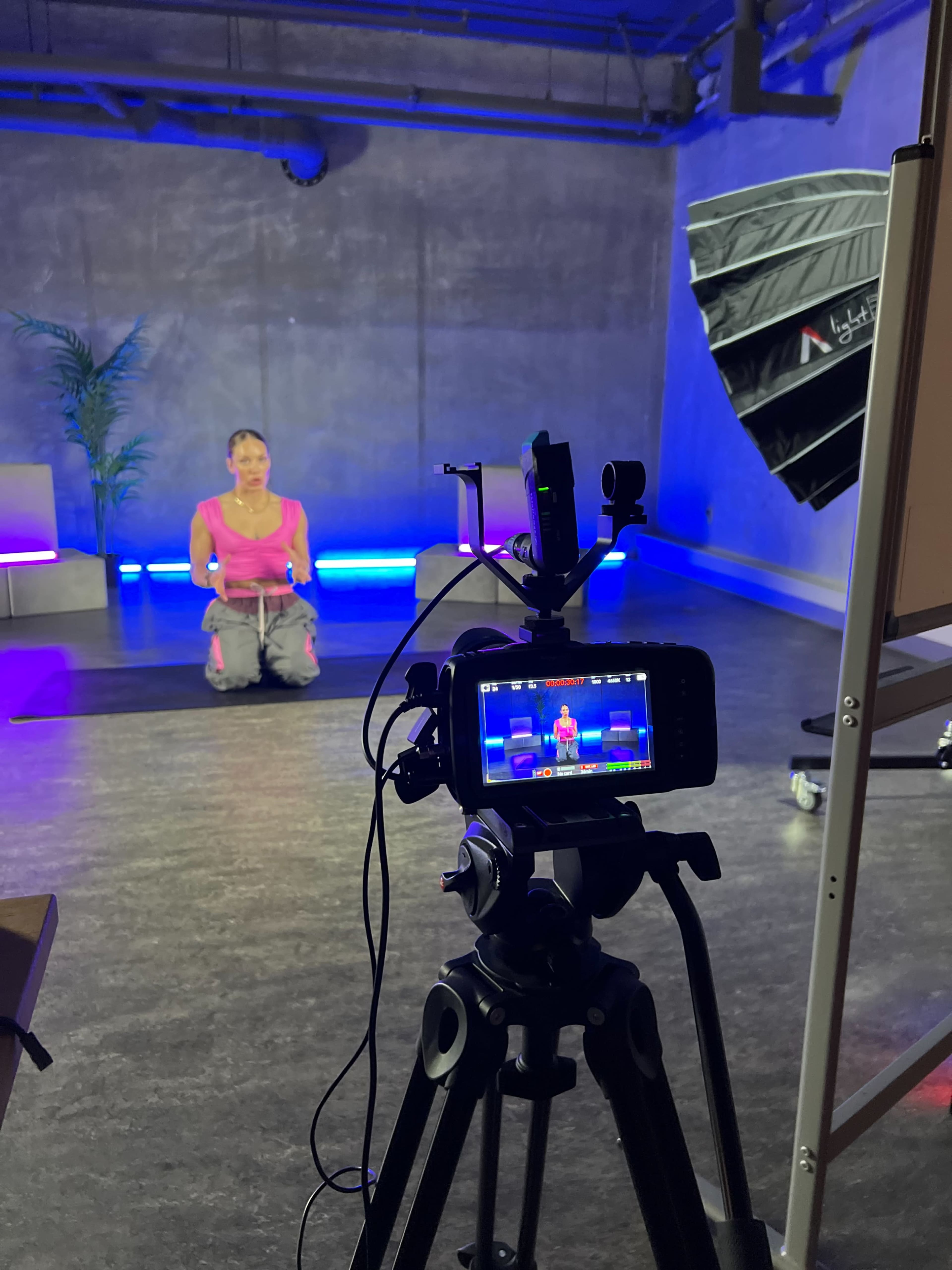 A woman is kneeling on a yoga mat in front of a camera setup with a bright light in a studio.
