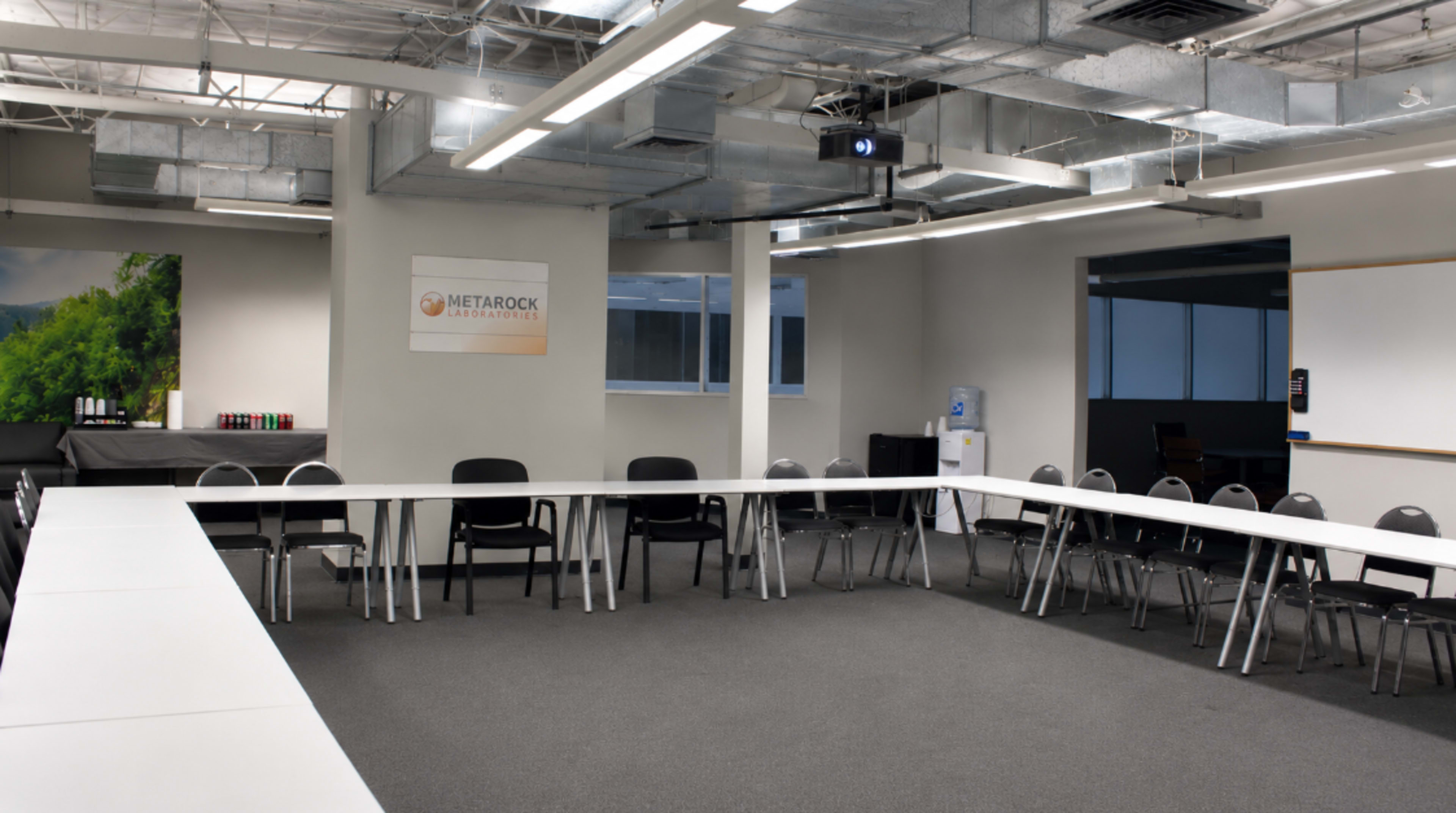 The image depicts a spacious conference room arranged with a long, rectangular table and chairs, featuring a projection screen and a water cooler in the background.