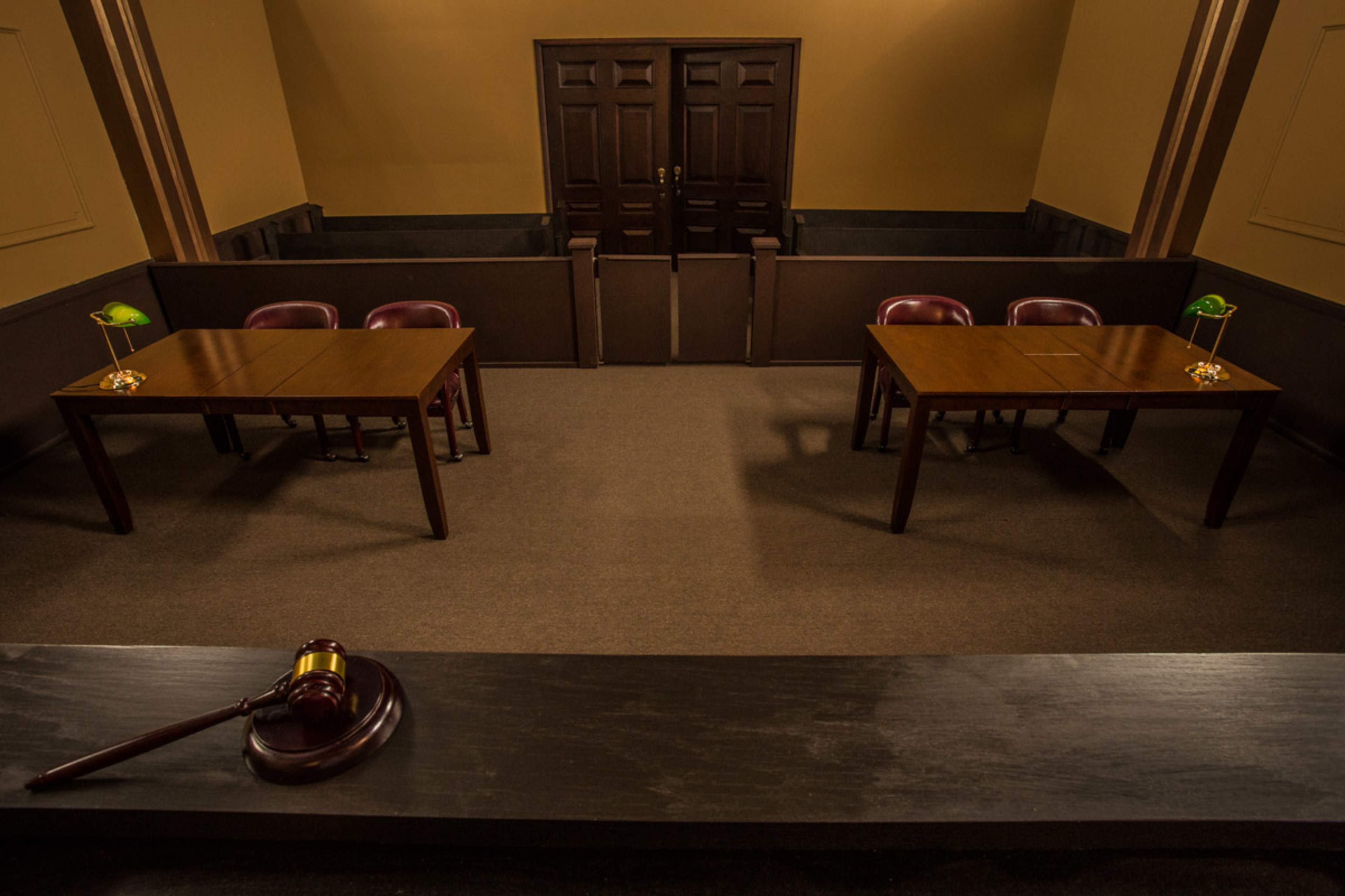 A courtroom interior features two wooden tables set for counsel, accompanied by green desk lamps, with a gavel resting on the front ledge.