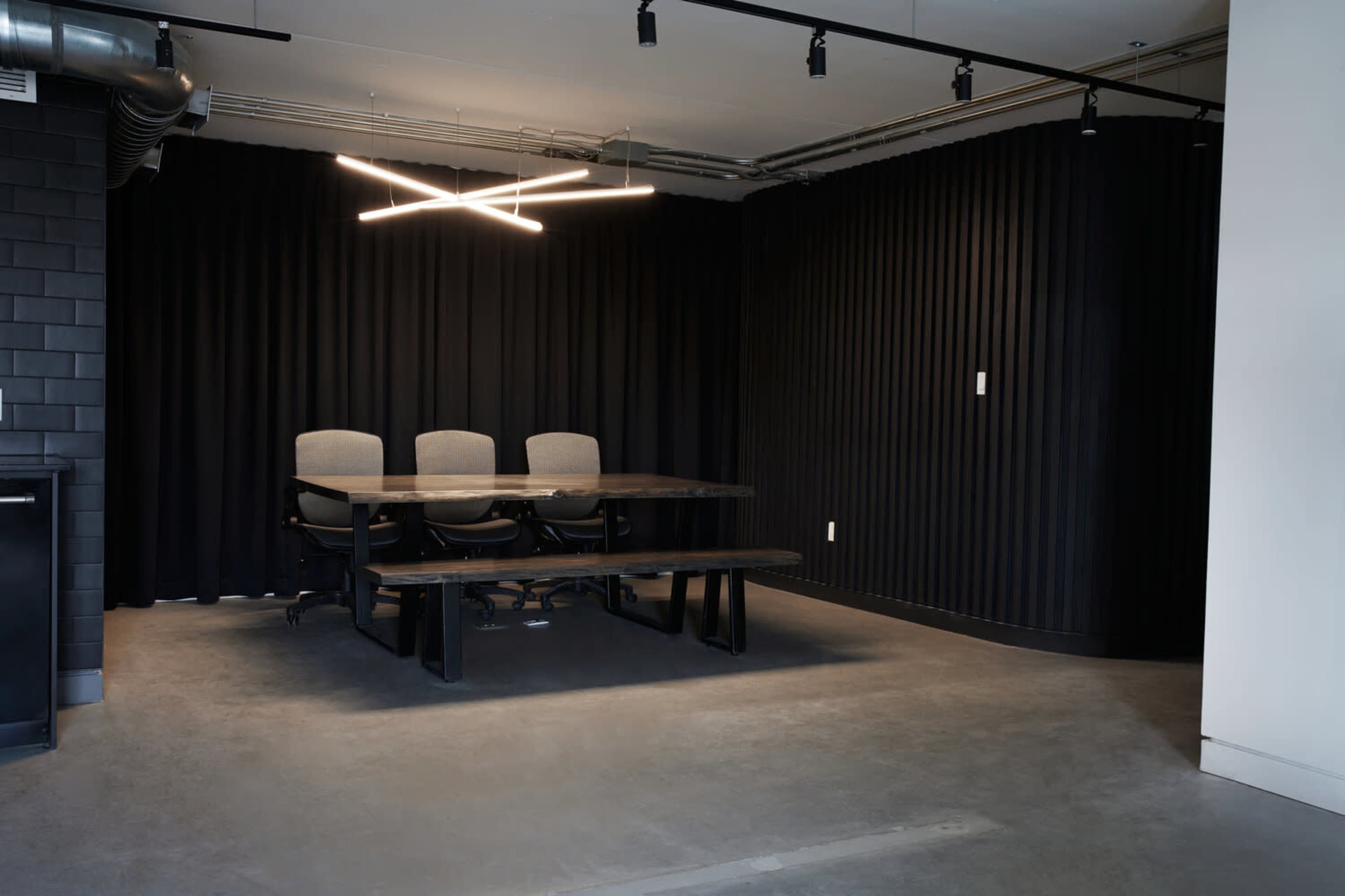 A conference room features a dark wall with three chairs and a long table under modern light fixtures.