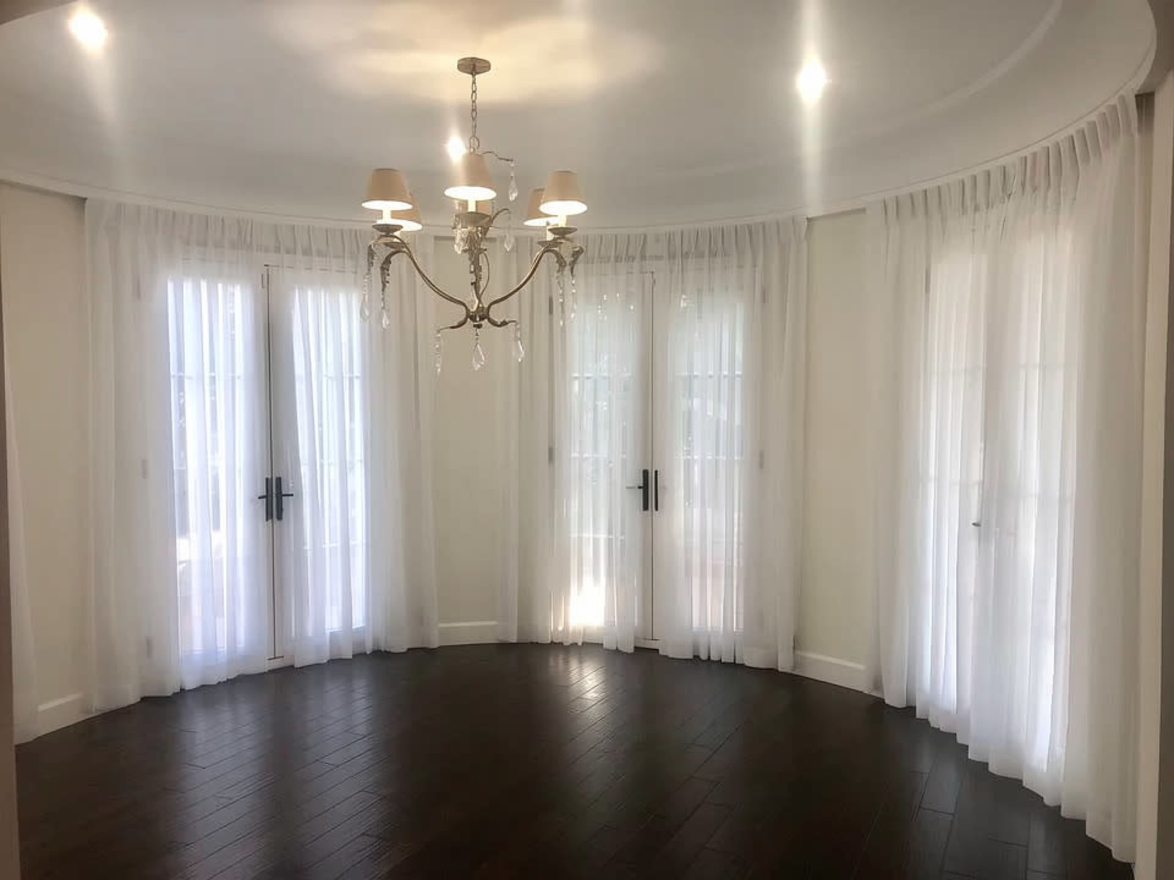 The image shows a curved room with wooden flooring, featuring large glass doors draped with sheer white curtains and a decorative chandelier hanging from the ceiling.