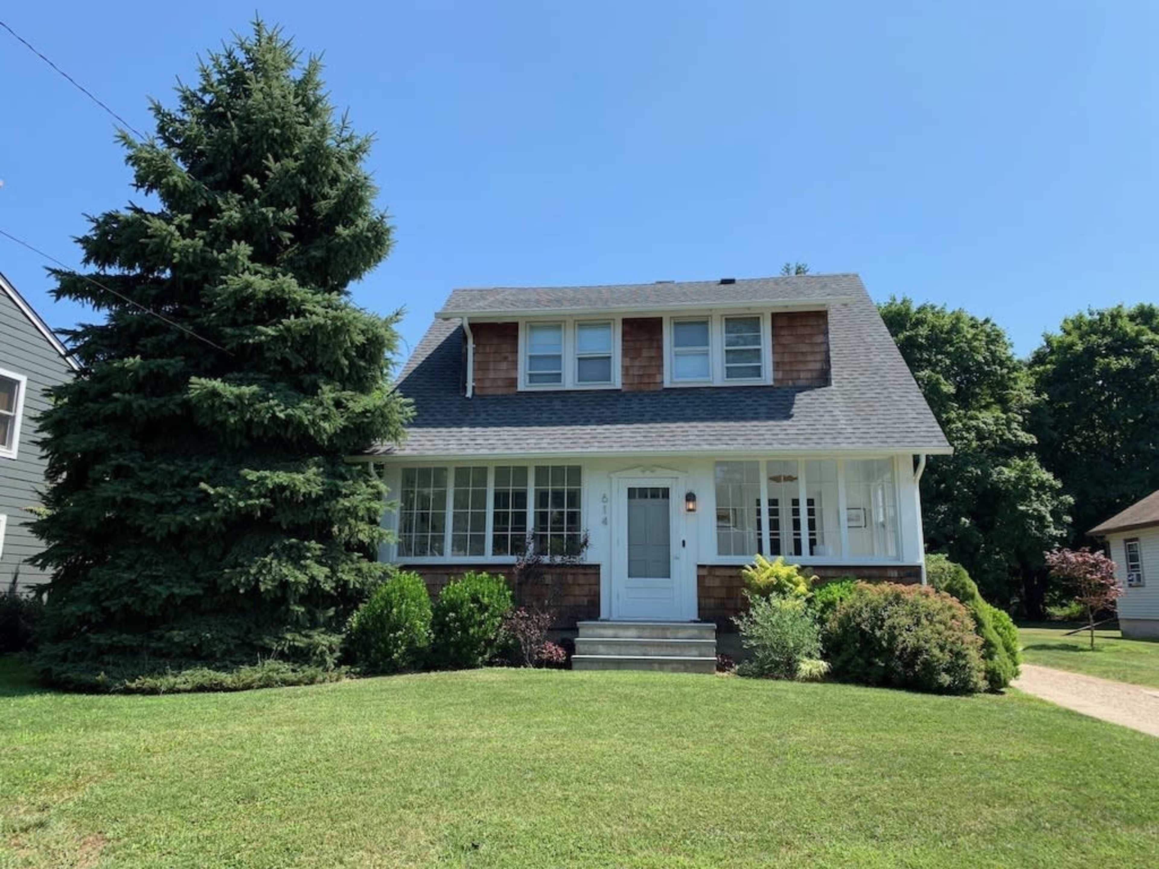 A two-story house with a gray roof and wooden siding sits on a grassy lawn surrounded by shrubs and trees.