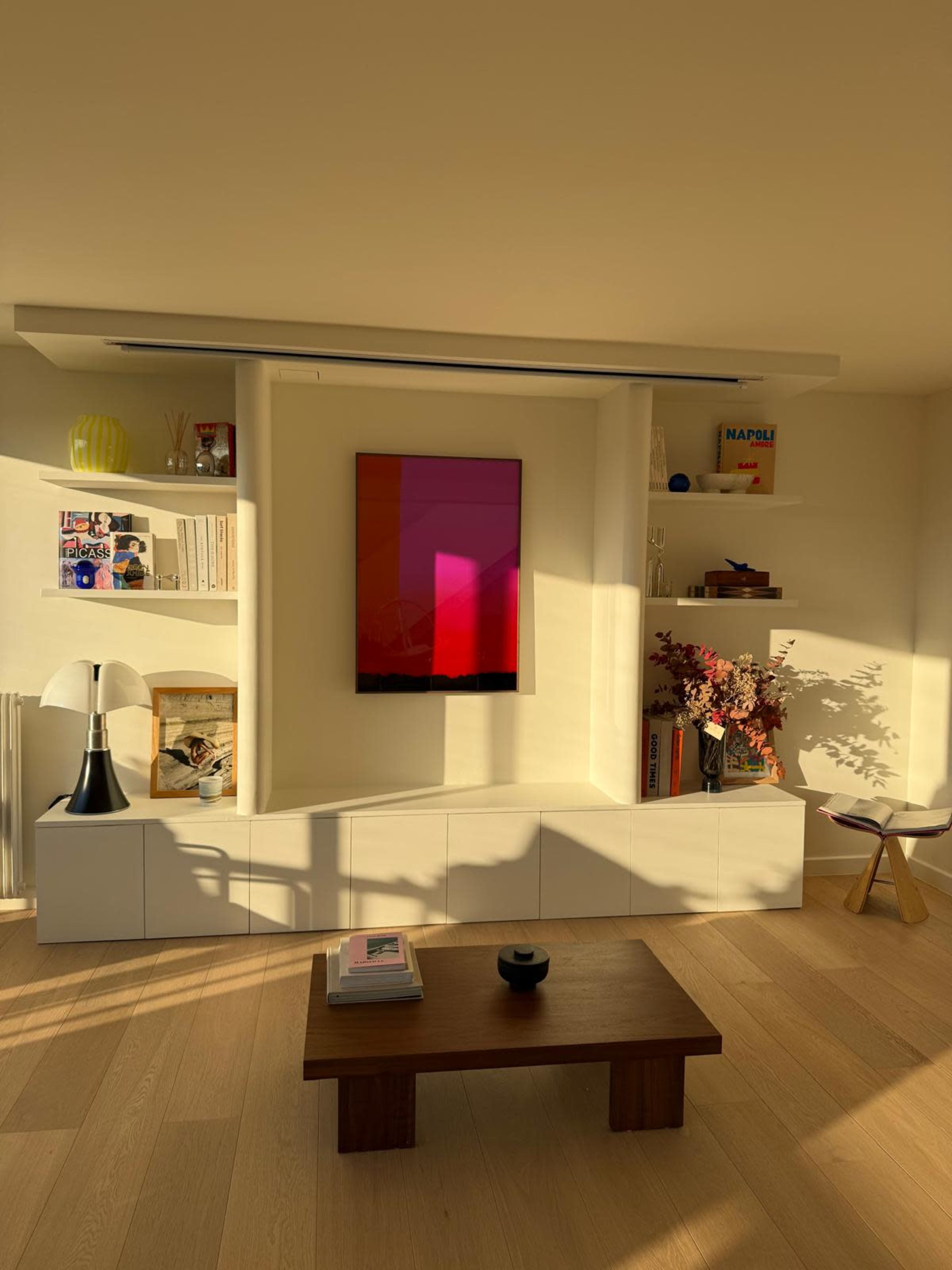 A modern living room features a sleek white entertainment unit, a wooden coffee table, and a wall-mounted artwork with a gradient design, bathed in warm sunlight.