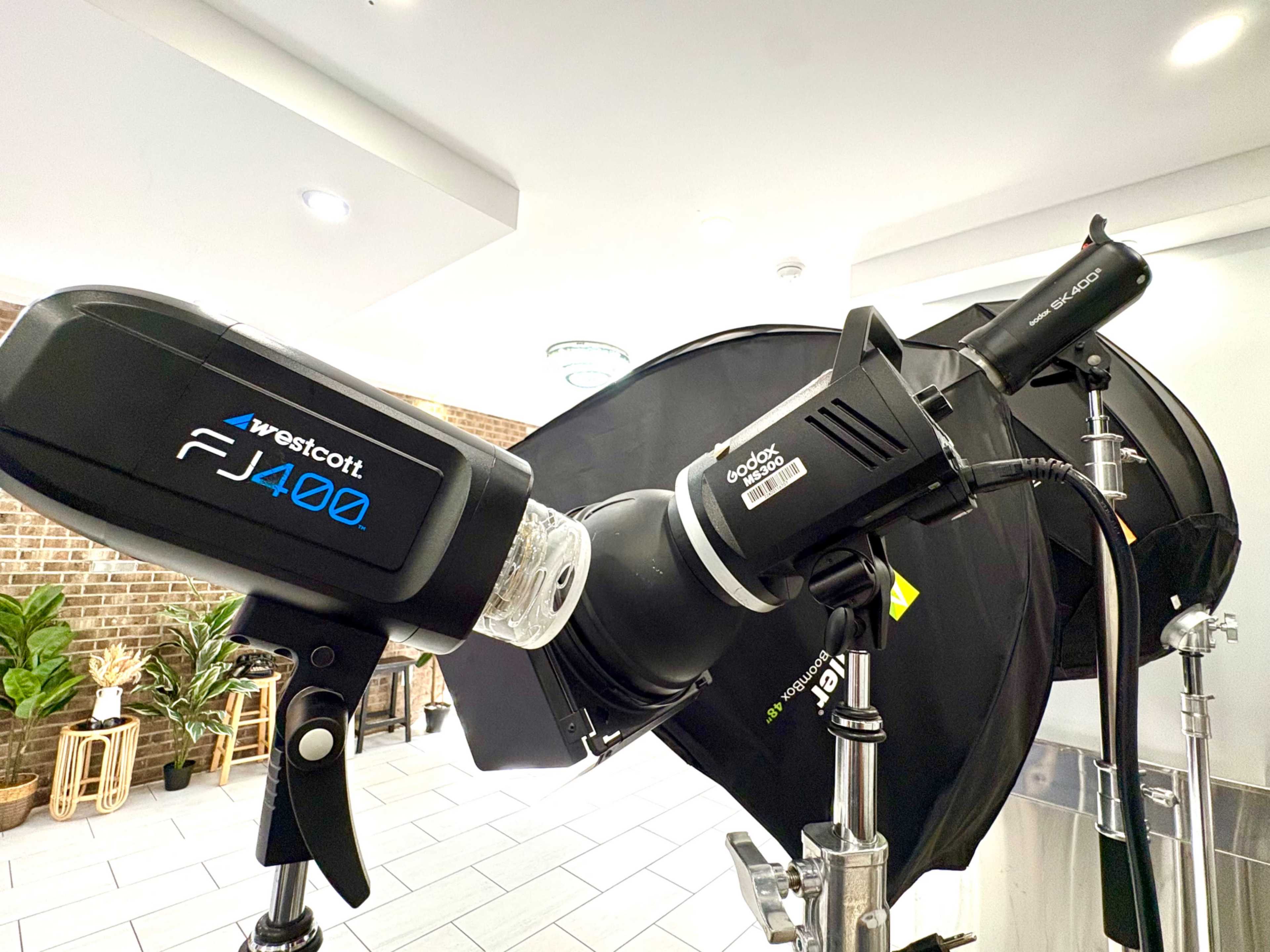 The image shows a Westcott FJ400 strobe light mounted on a stand, positioned next to a black softbox, with brick walls and potted plants in the background.