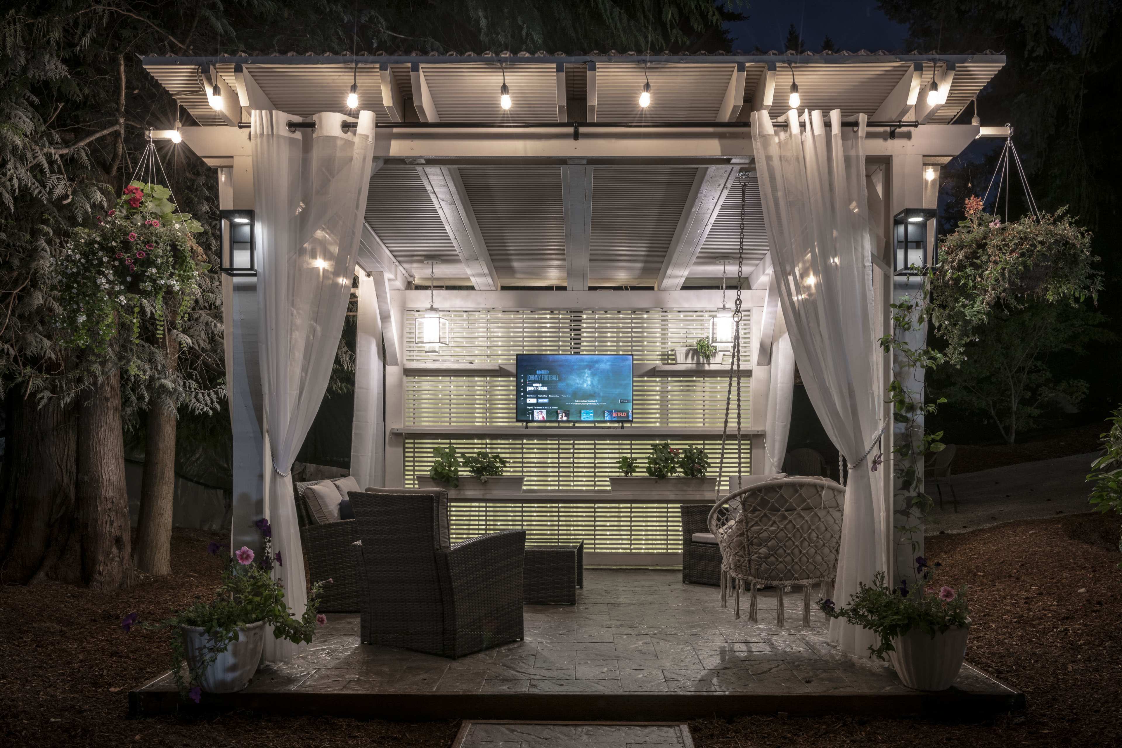 A covered outdoor gazebo features a television mounted on a wall, surrounded by seating and decorative plants, illuminated by hanging lights.