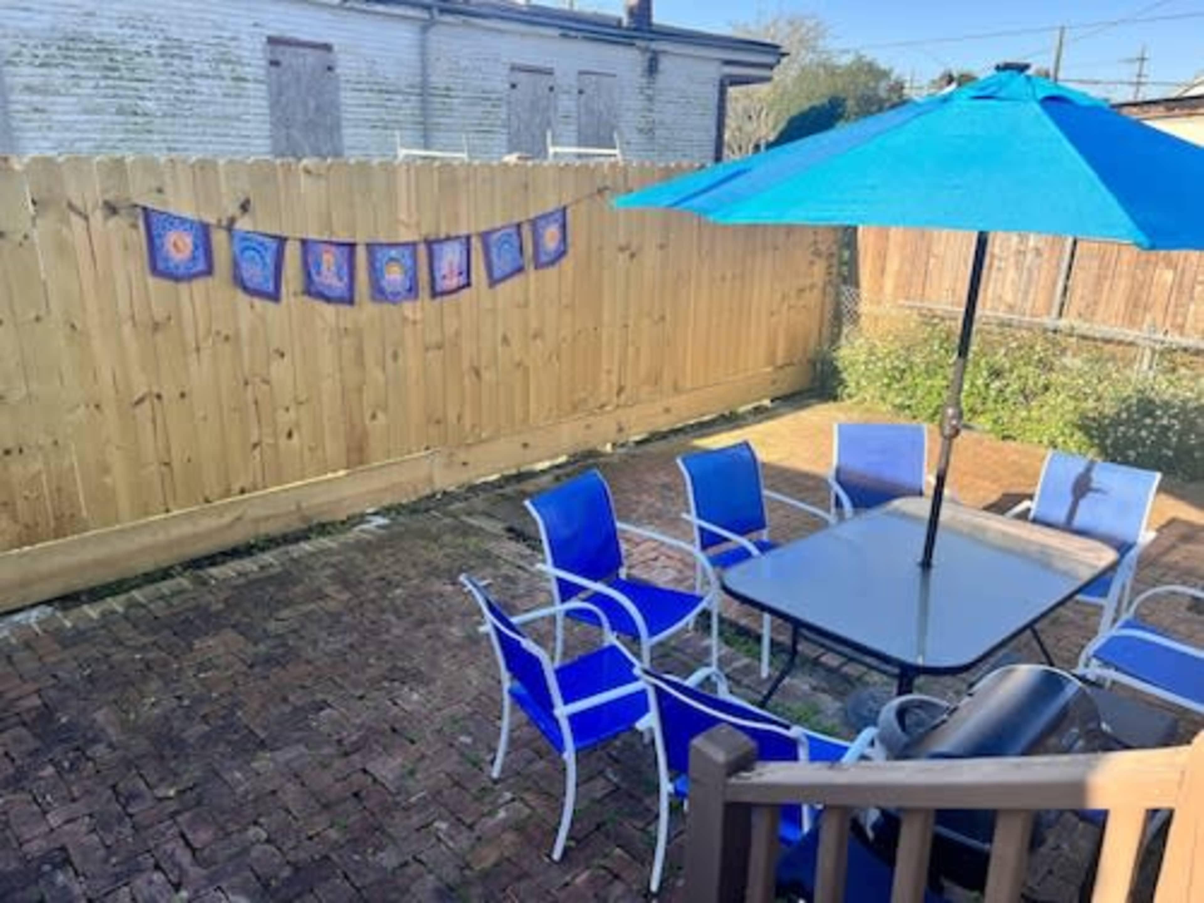 A backyard patio features a blue umbrella over a black table surrounded by blue chairs, with a wooden fence displaying a line of colorful decorations.