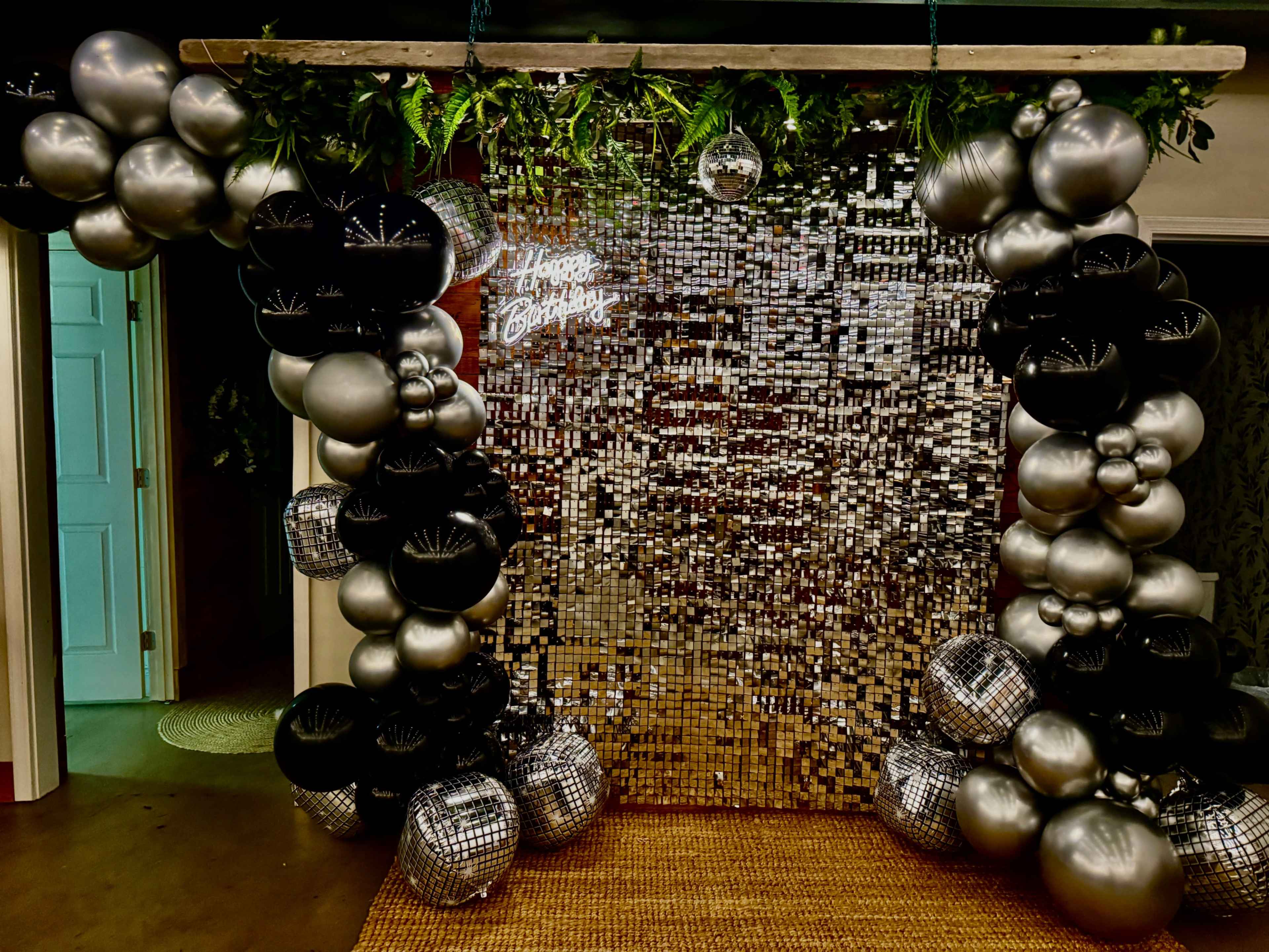 A decorative arch made of black and silver balloons frames a sparkling mirrored backdrop with a glowing "Happy Birthday" sign.