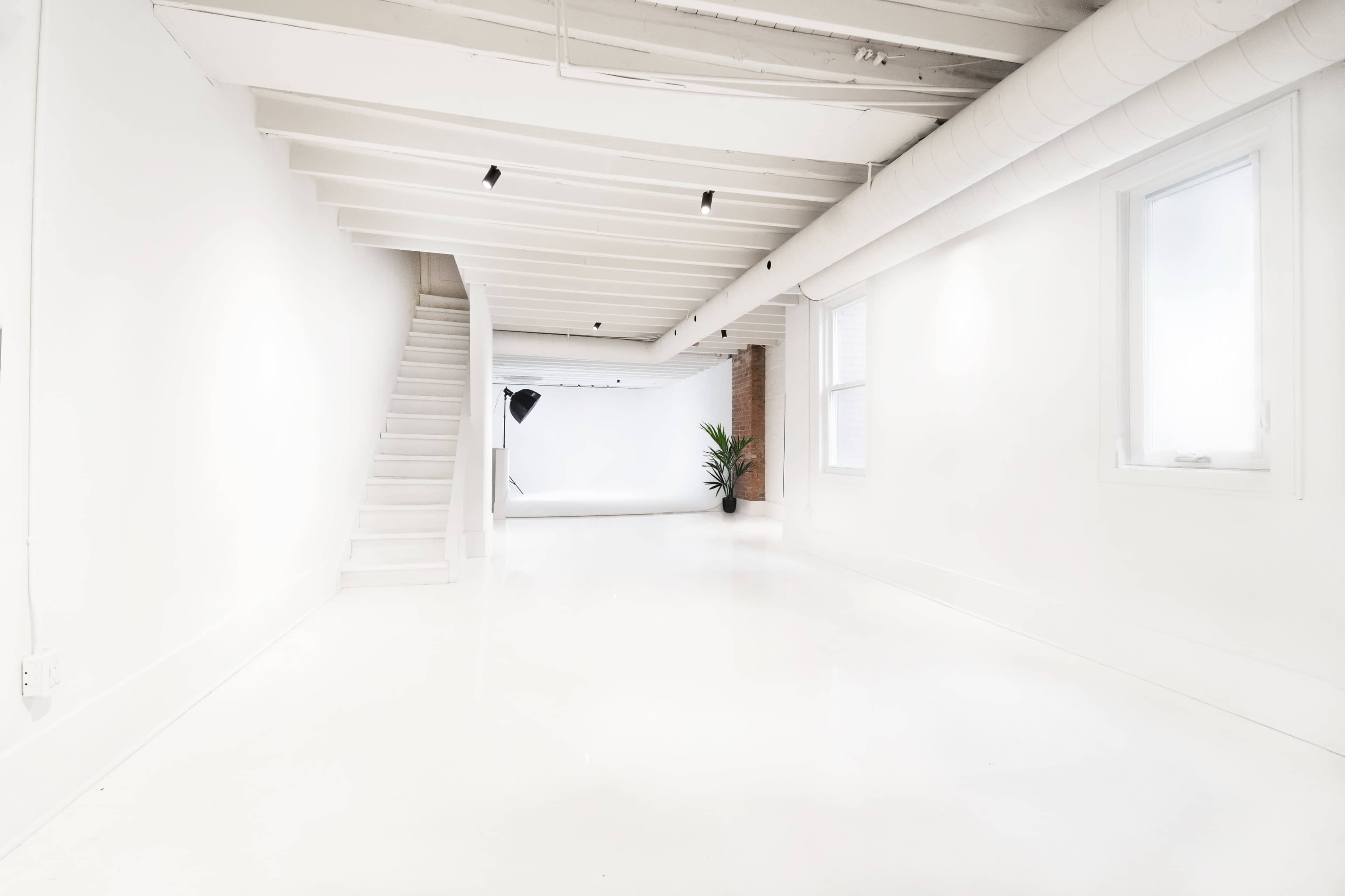 A bright, empty studio space with white walls and flooring, featuring a staircase on the left and a backdrop with a potted plant on the right.