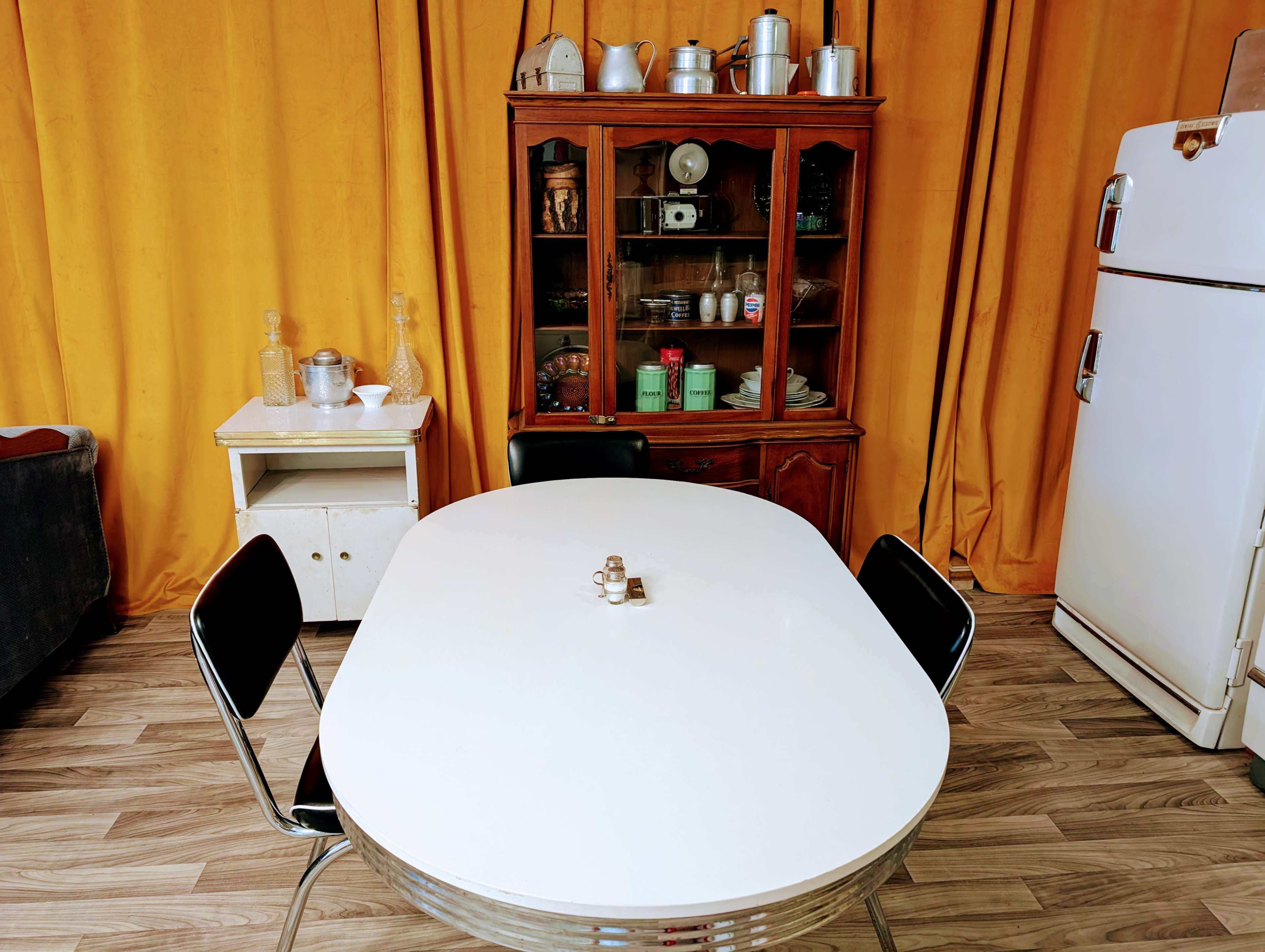 A white oval dining table is set in a room with orange curtains, surrounded by black chairs, with a wooden cabinet displaying glassware and kitchen items in the background.